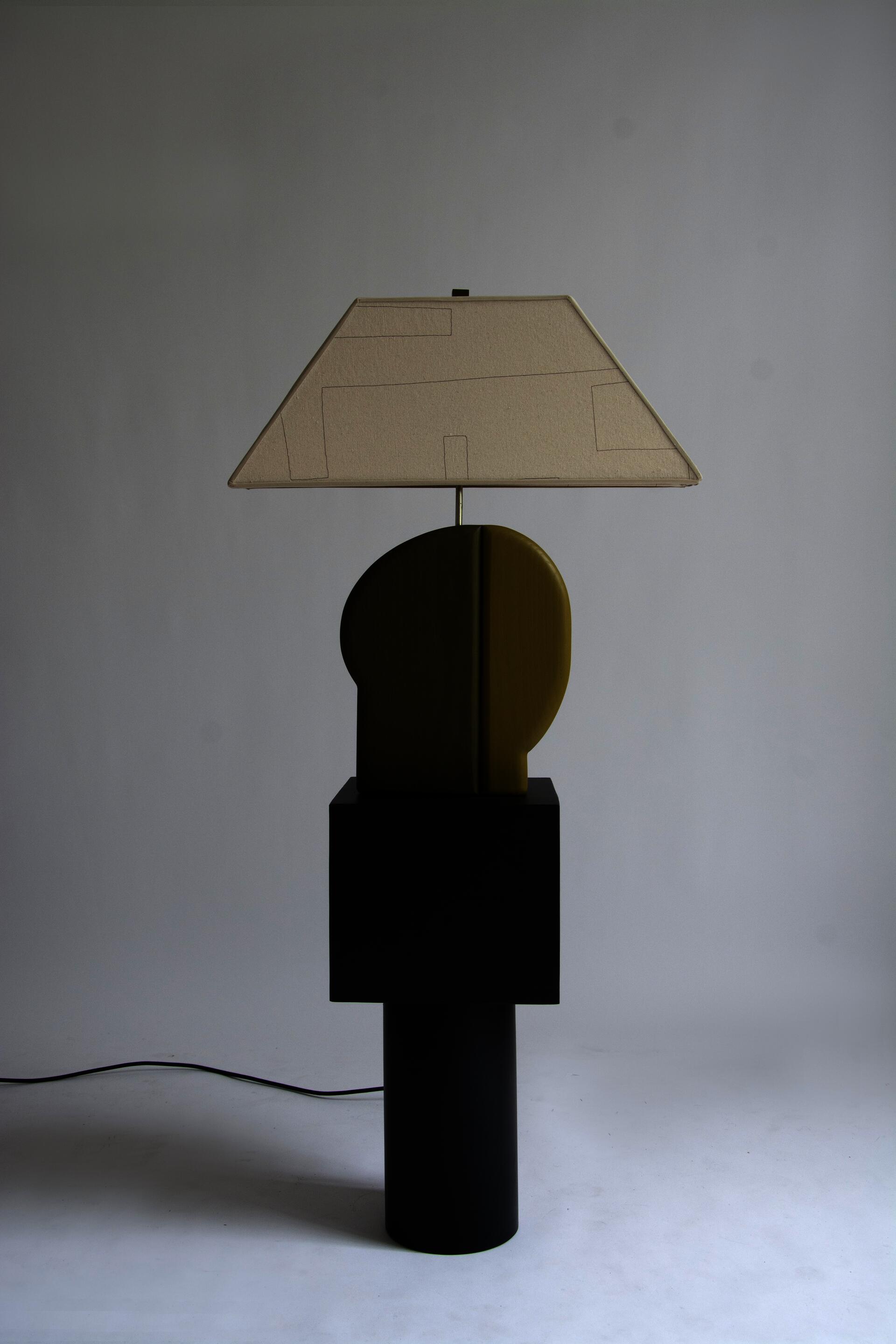 The Side table has a black wooden cylindrical base, with a square black wooden top. A two toned green wooden lamp base and canvas shade with hand sewn lines sits atop the table.