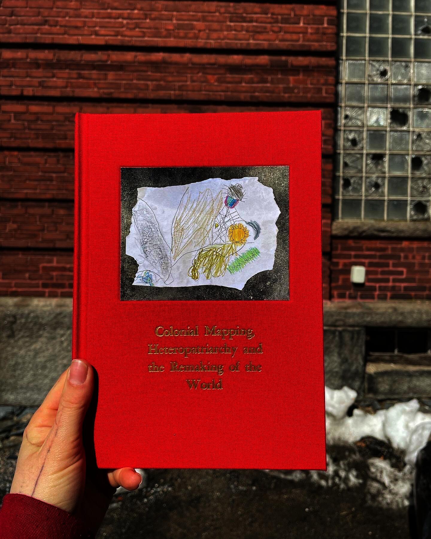 A hand holding a red book titled, "Colonial Mapping, Heteropatriarchy, and the Remaking of the World", with an image of a drawing.