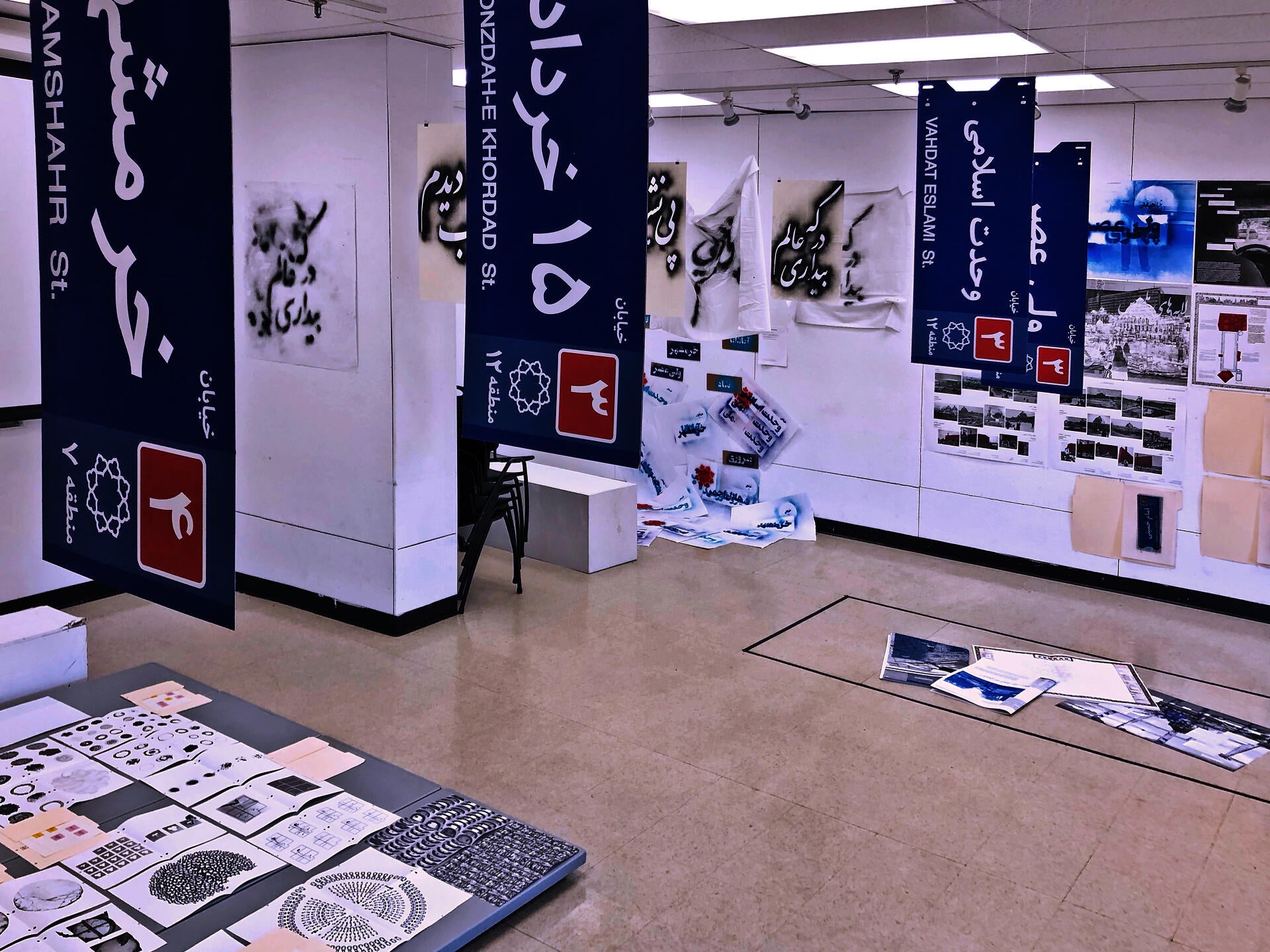 This image shows a multi-component installation in a room with objects hanging from the ceiling, printed outs pinned on the wall, and books laid on the ground