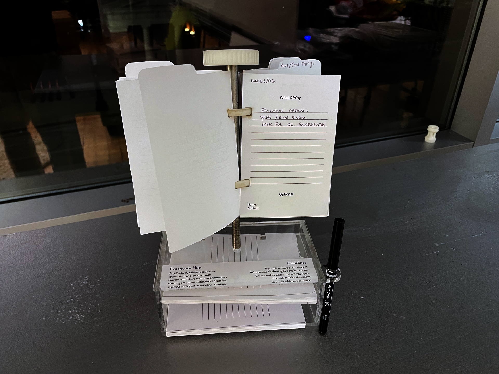 A small steel pole holding half sheets of paper that people have written on. The pole is on top of an clear box holding extra paper and a pen.
