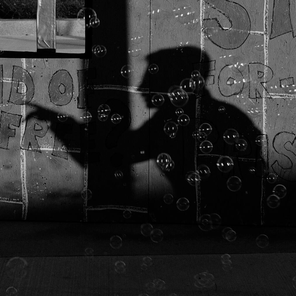 the shape of a shadow pointing somewhere out of frame, bubbles bubbling floating there is cardboard behind the pointing shadow, these words (or partial words) written by hand onto the cardboard: "free?" "for -" "pris -"