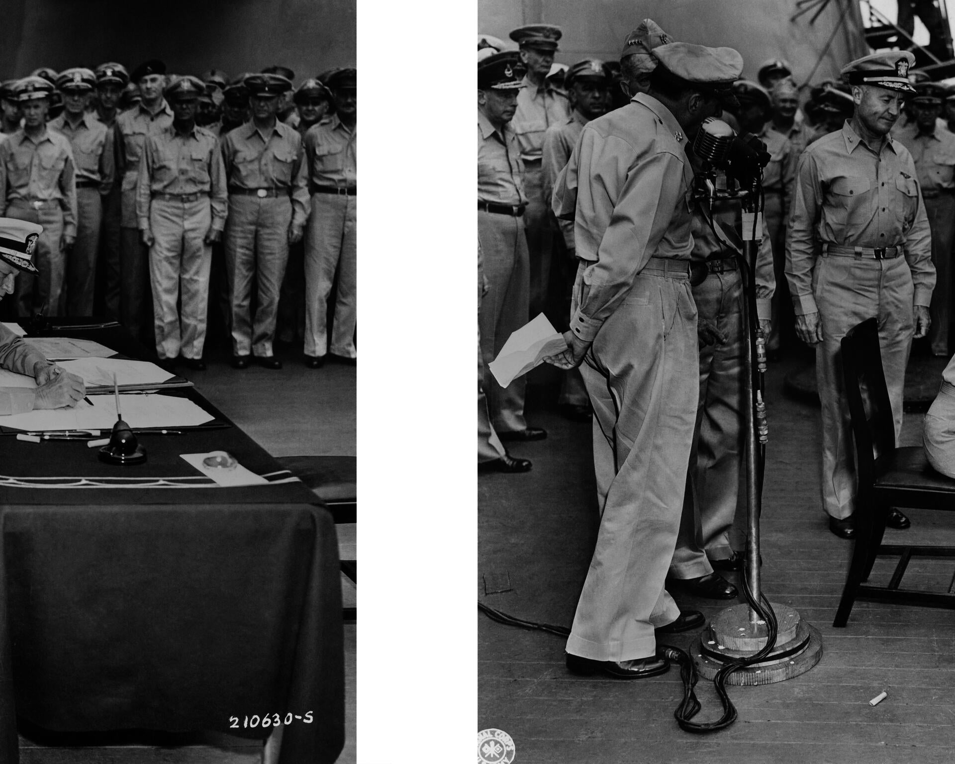 A black-and-white photograph of a crowd of soldiers looking on as a figure–cropped out of the frame–signs the document that officiates Japan's surrender at the close of World War II.