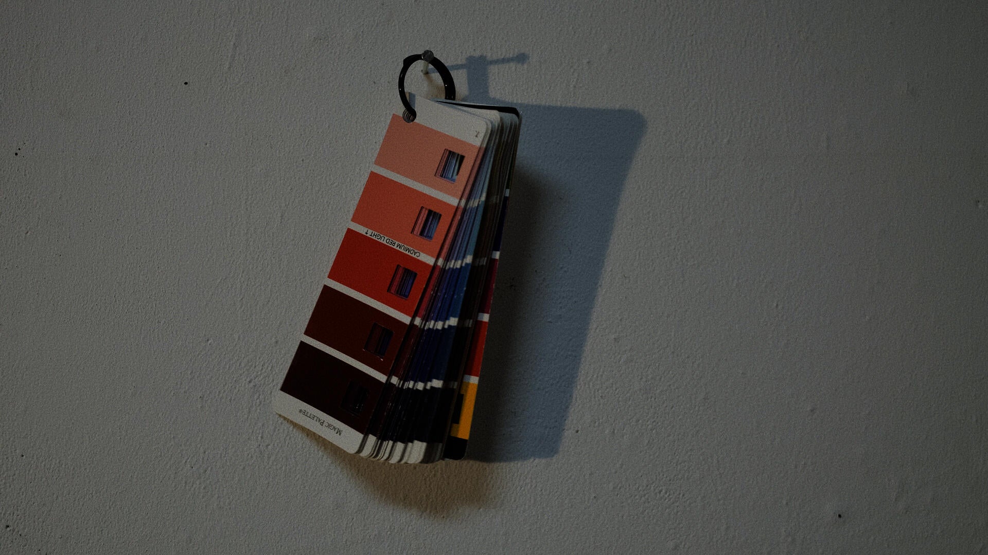 A color index card showing different shades of red is nailed to a white wall.