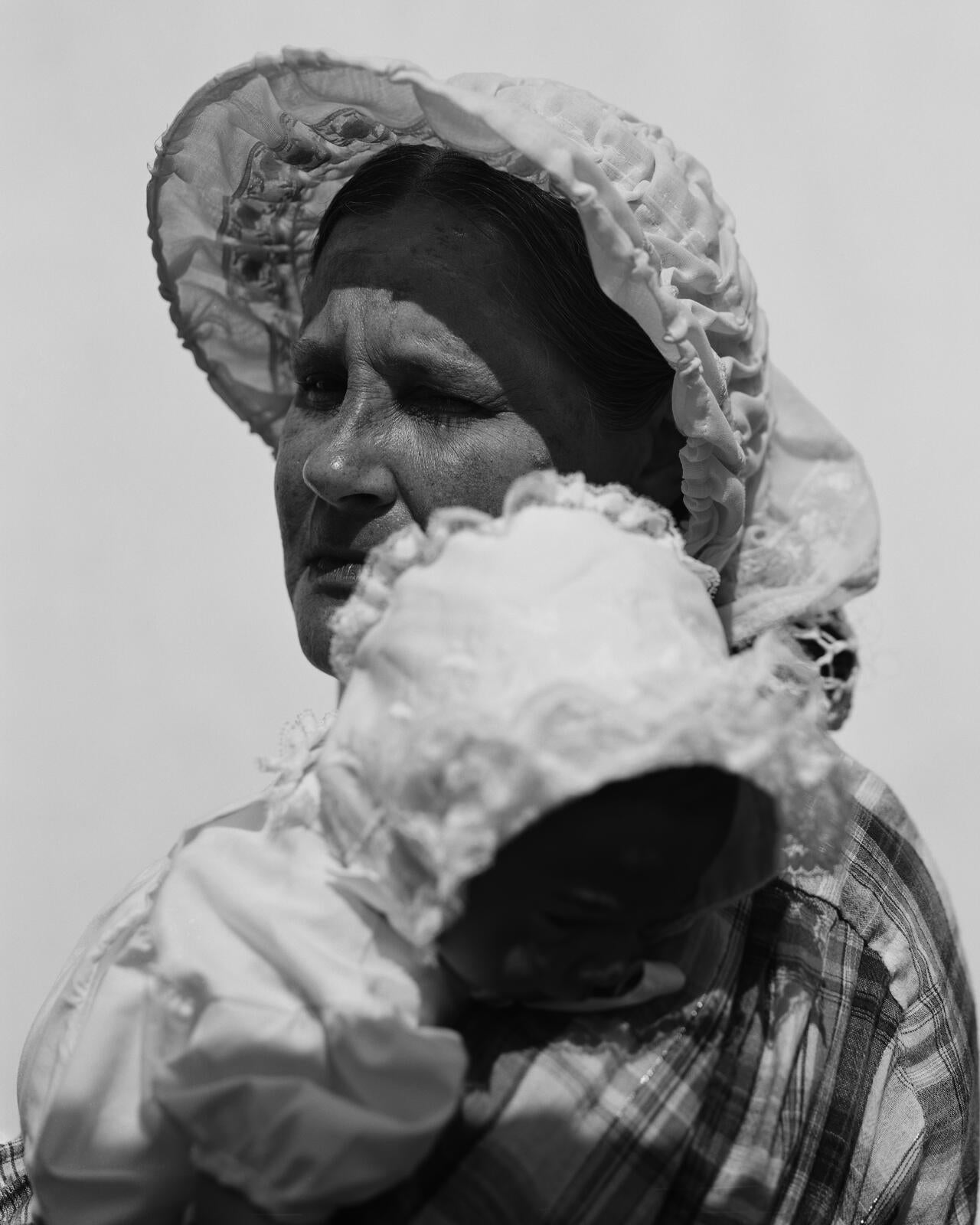 A woman in 19th century clothes stands against a white backdrop in the bright sun. She looks at the camera with one open eye. She is carrying a baby in her arms.