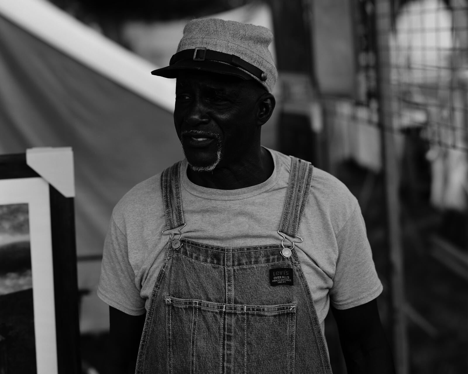 A Black man stands in the center of the frame looking off to the left, he is wearing a Confederate hat. Behind him are canvases and frames and posters.