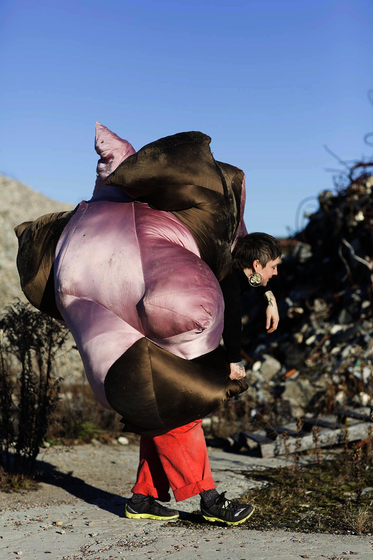 A person with short salt and pepper hair wearing red pants carries an immense and strangely shaped pillow-like object amongst pieces of tattered rebar and broken steel.