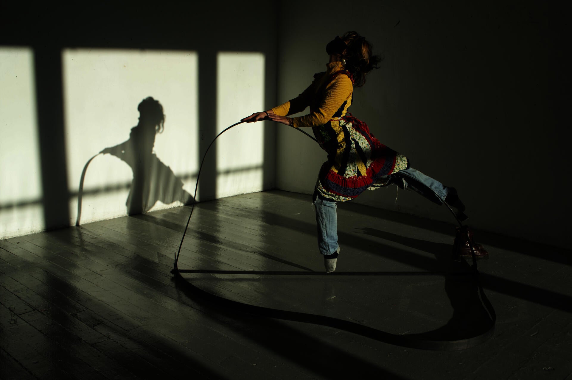 A person with dark hair and a colorful dress layered over jeans balances and dances atop a delicately bent steel beam.