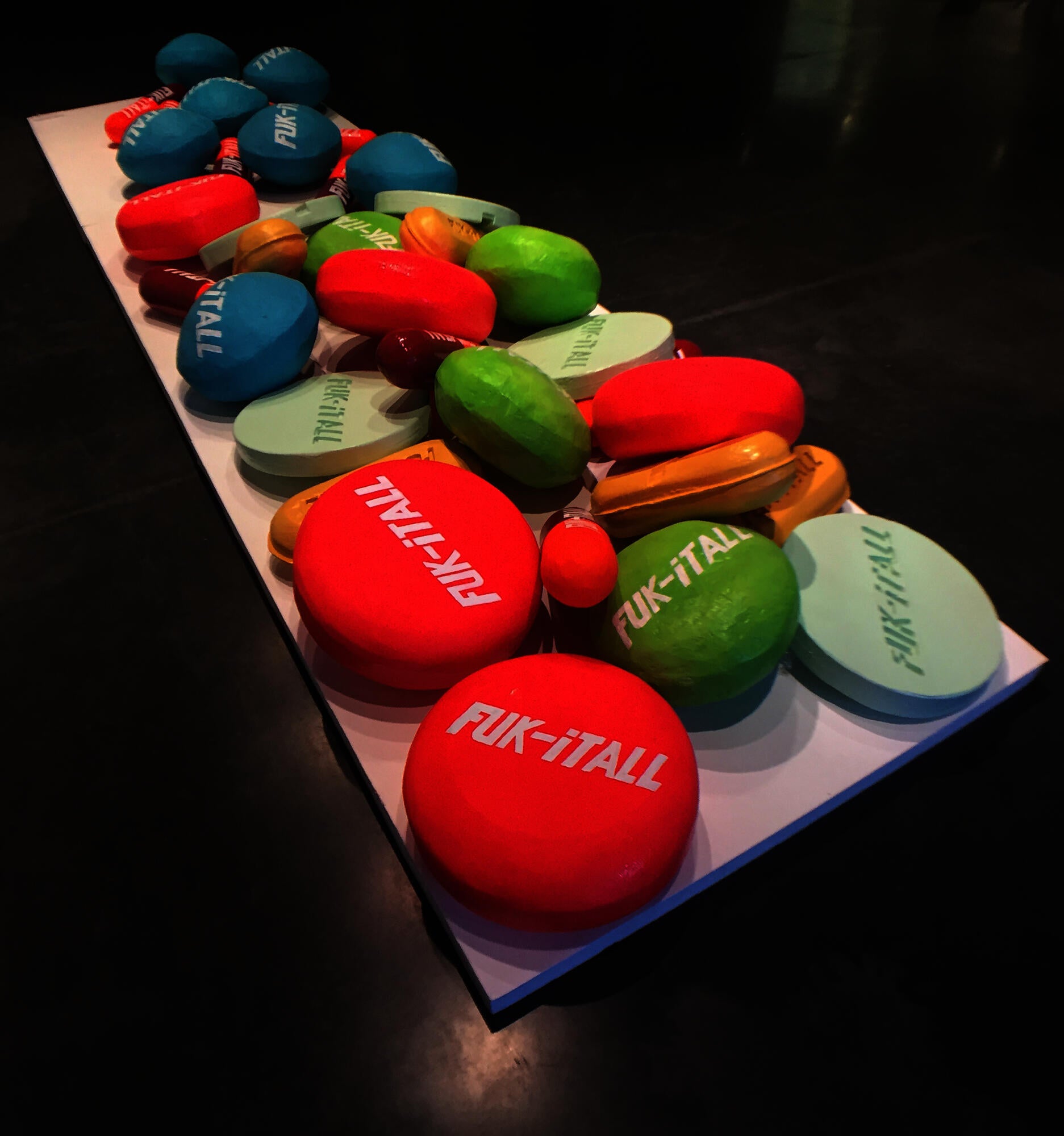 The work is an assortment of brightly colored basketball-sized pills which are of varying lengths, shapes, and designs. These pills are placed on a stretched-out platform that takes up a prominent area in the center of the floor. It is made of mixed media of rubber and wood on foam board.