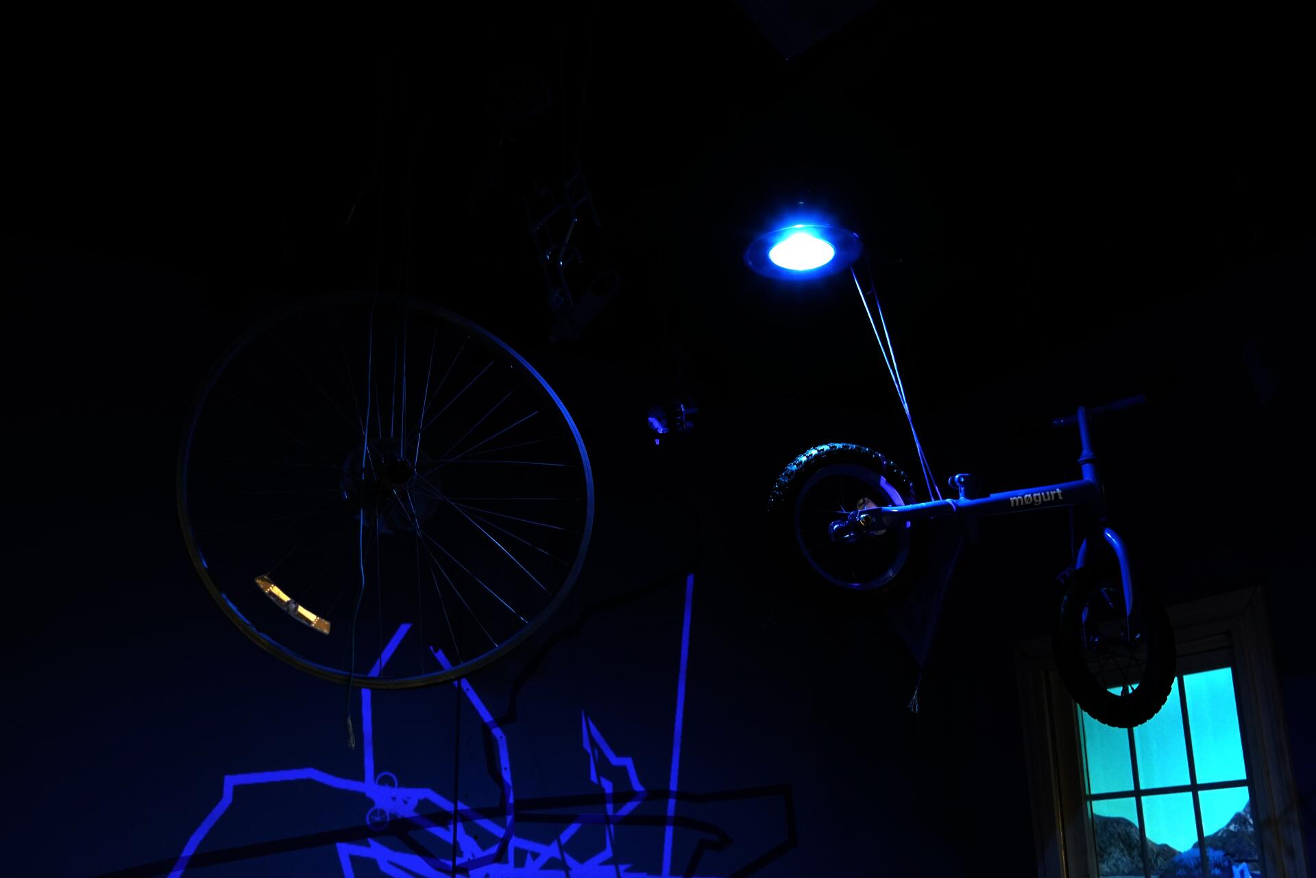 Installation materials suspended from the ceiling: Mouse trap board game pieces, kid's bike, busted bike wheel, paracord