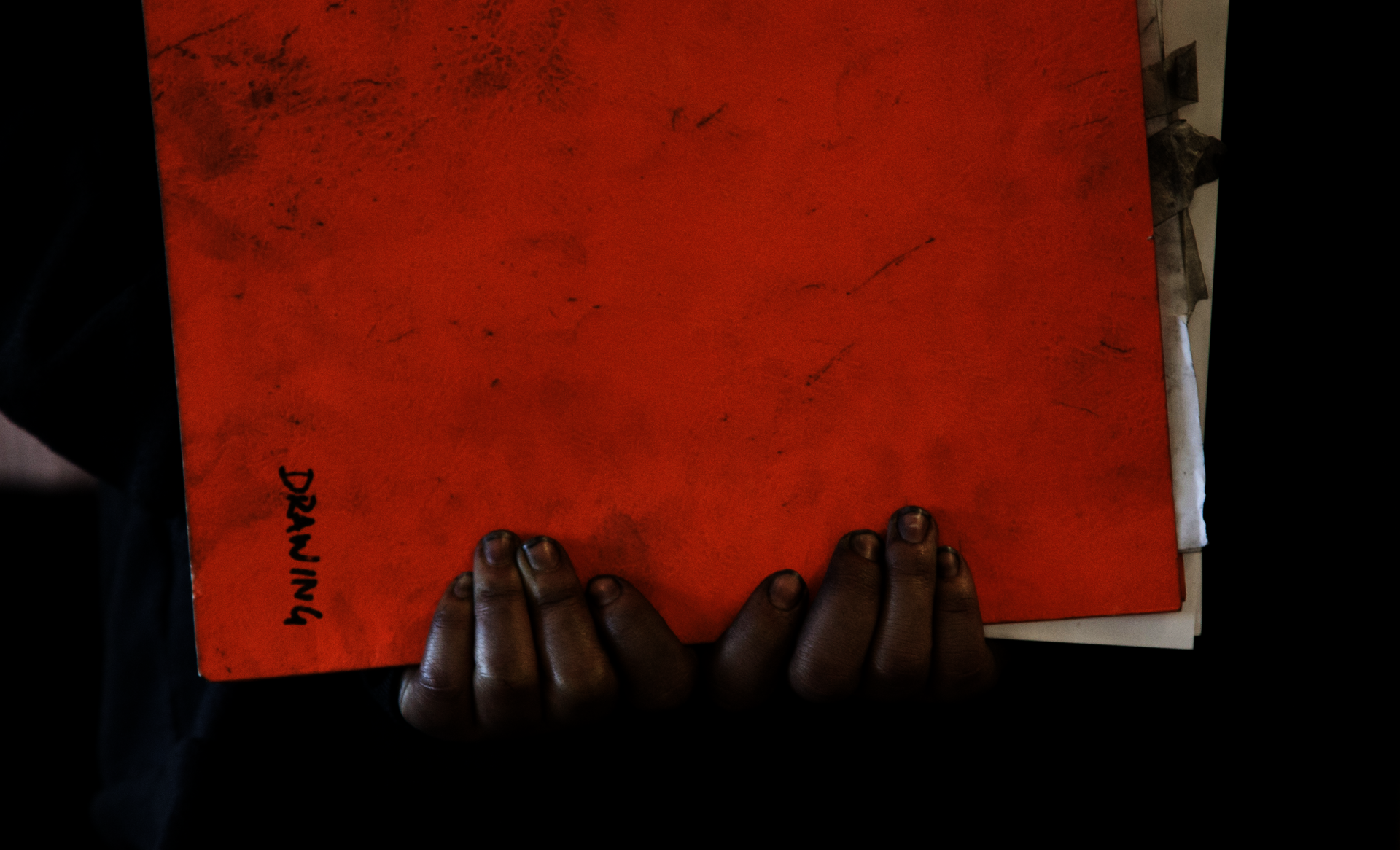 A pair of young Black hands grasp an orange folder labeled "Drawing"