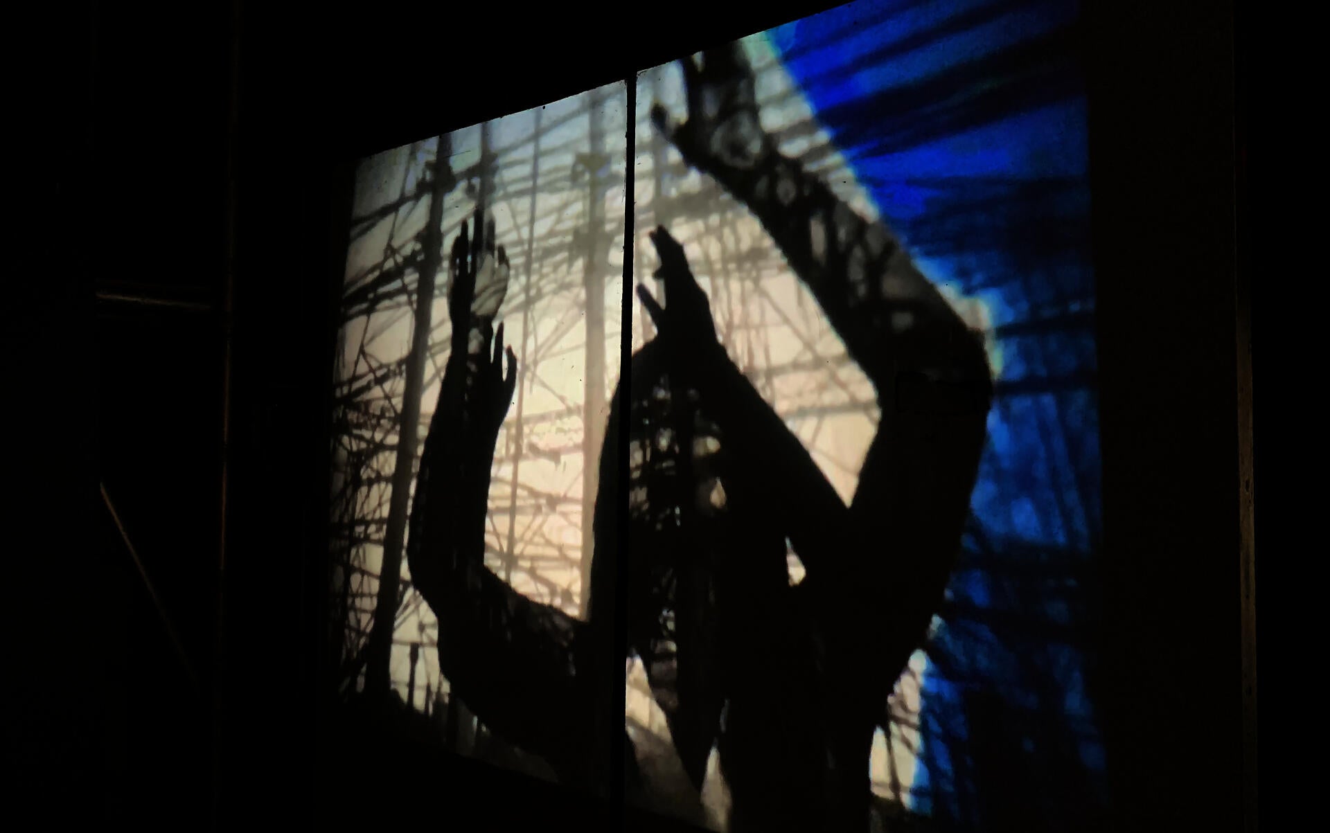 Frame from an animation lights up a dark room. It shows a human figure reaching amidst a web of lines like scaffolding or telephone poles and lines