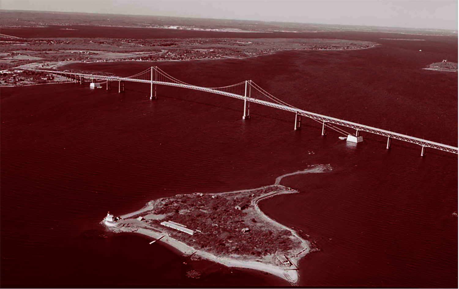 red-tinted aerial view of the Pell Bridge, a suspension bridge spanning a river with an island