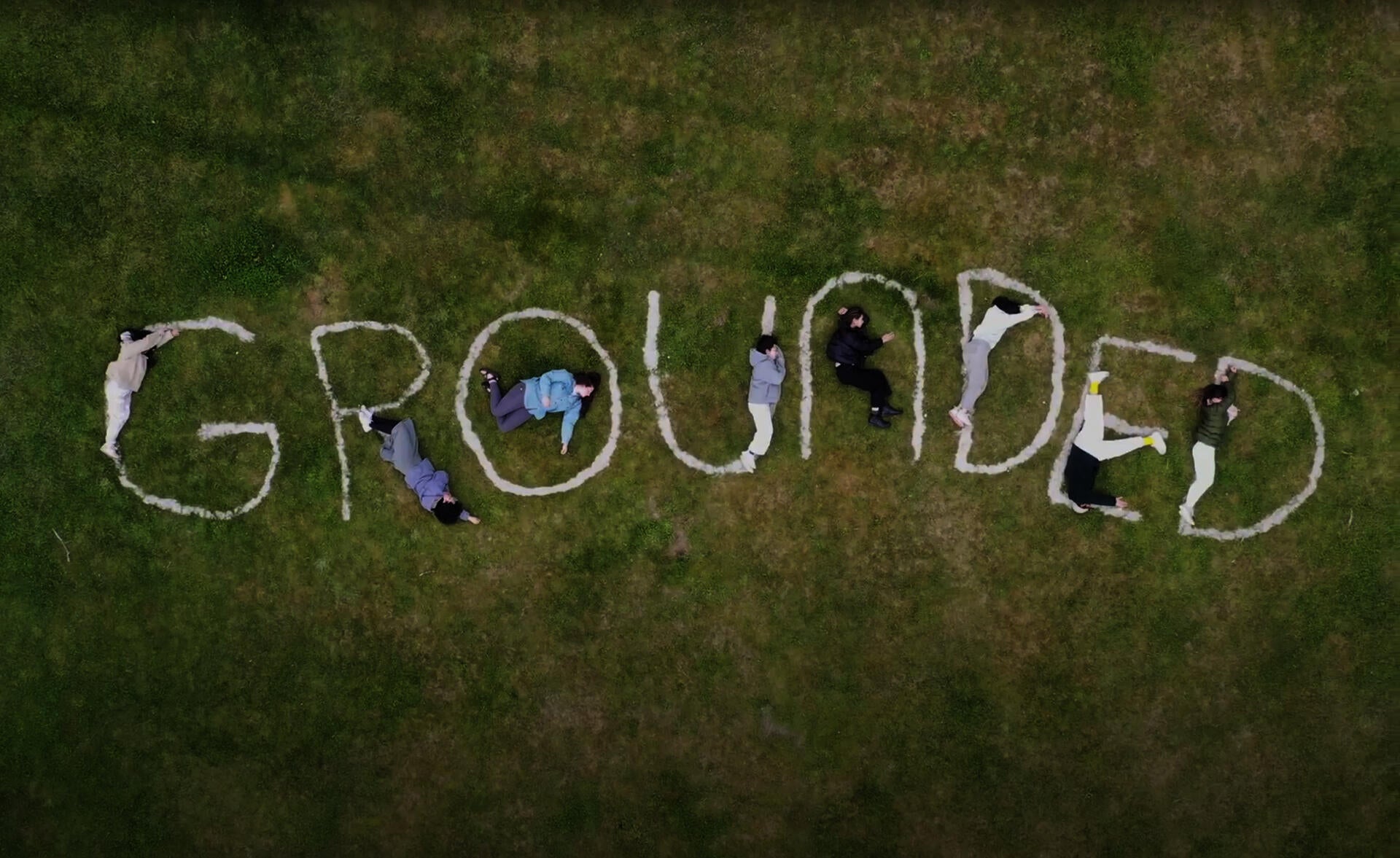 Aerial photo of a field of grass with the word "grounded" spelled out. Human figures lay on the ground, interacting with the letters.