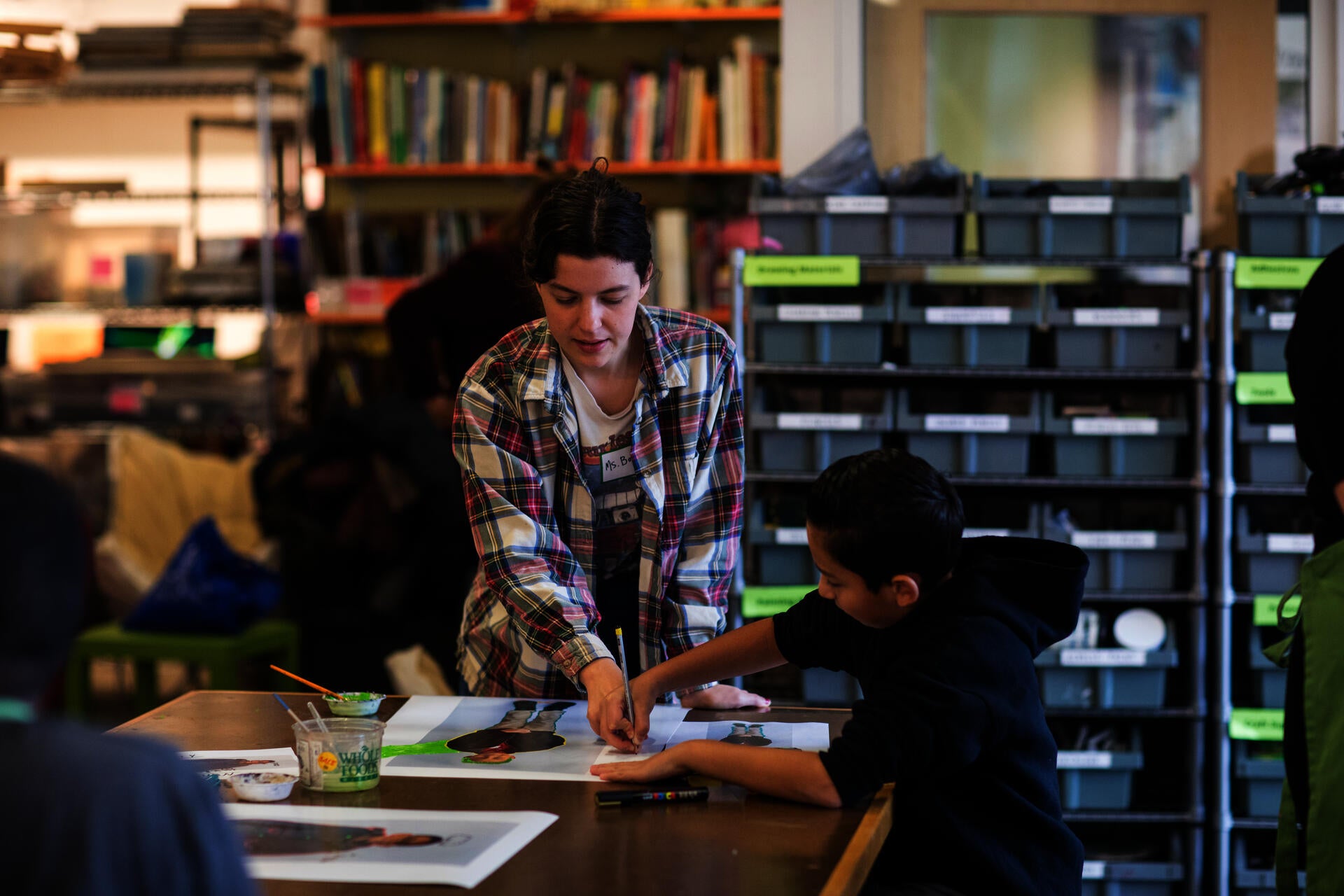 In a classroom setting, a young white woman assists a Latinx child holding a pencil as he draws on a photograph.