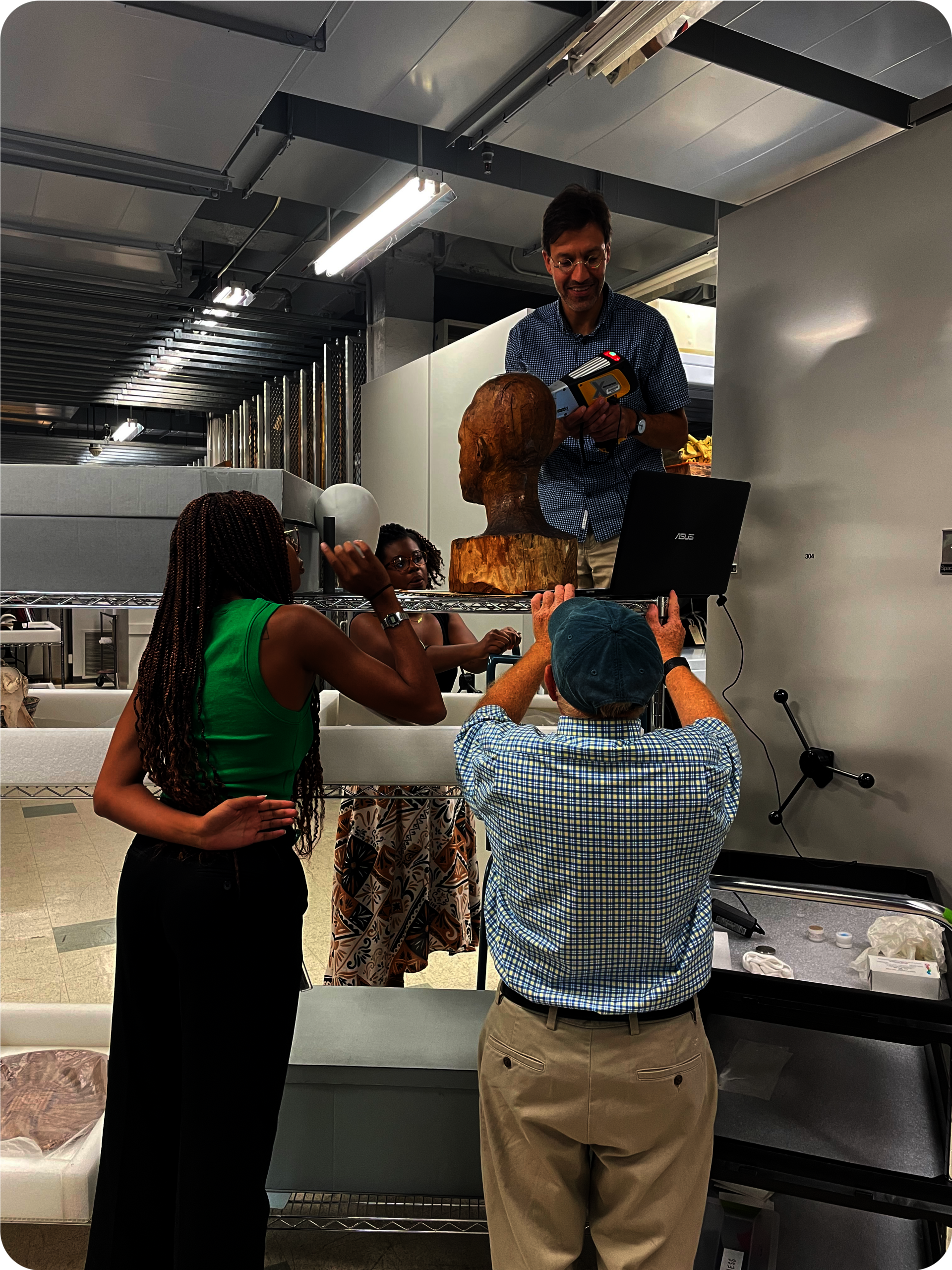 Four people around a wooden bust next to a laptop. Two people raise their arms towards the bust. Another looks sideways, while another standing on a platform holds a tool.