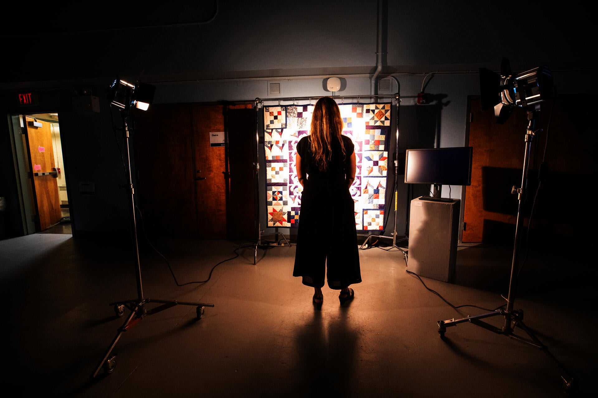 A woman stands in a classroom, in front of a suspended quilt lit by two powerful fresnel lights with barn doors.