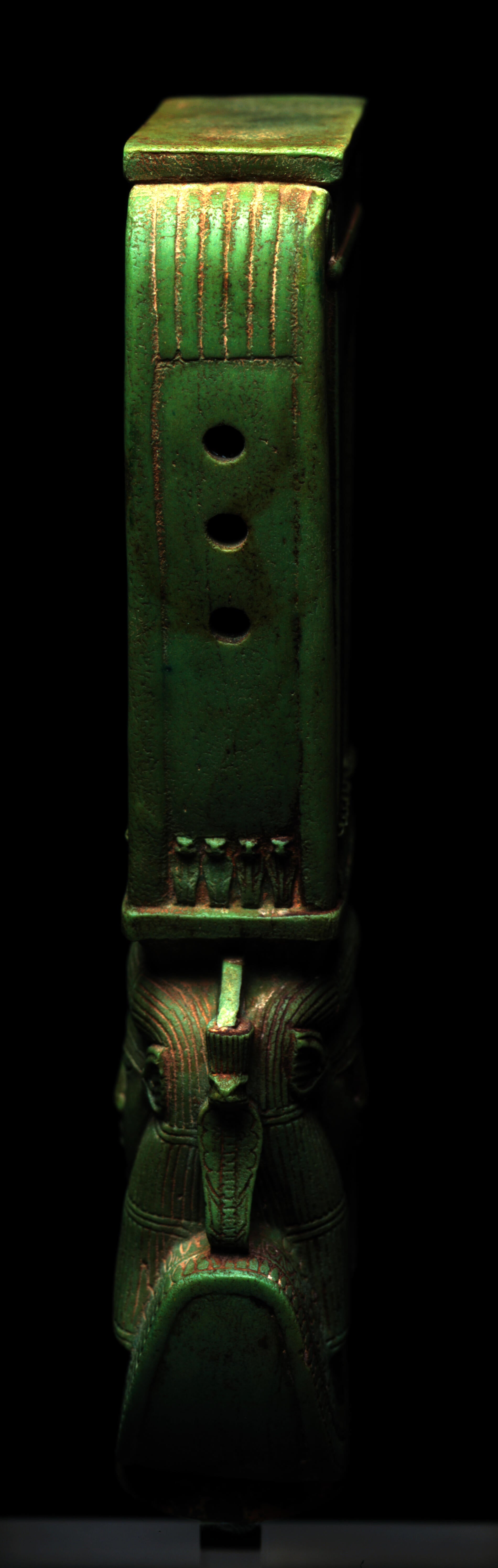 Side-view of a long jade-green sculpture depicting a collared figure’s head wearing a tall headdress. The headdress has three holds in the center and small snake figurines at the base.