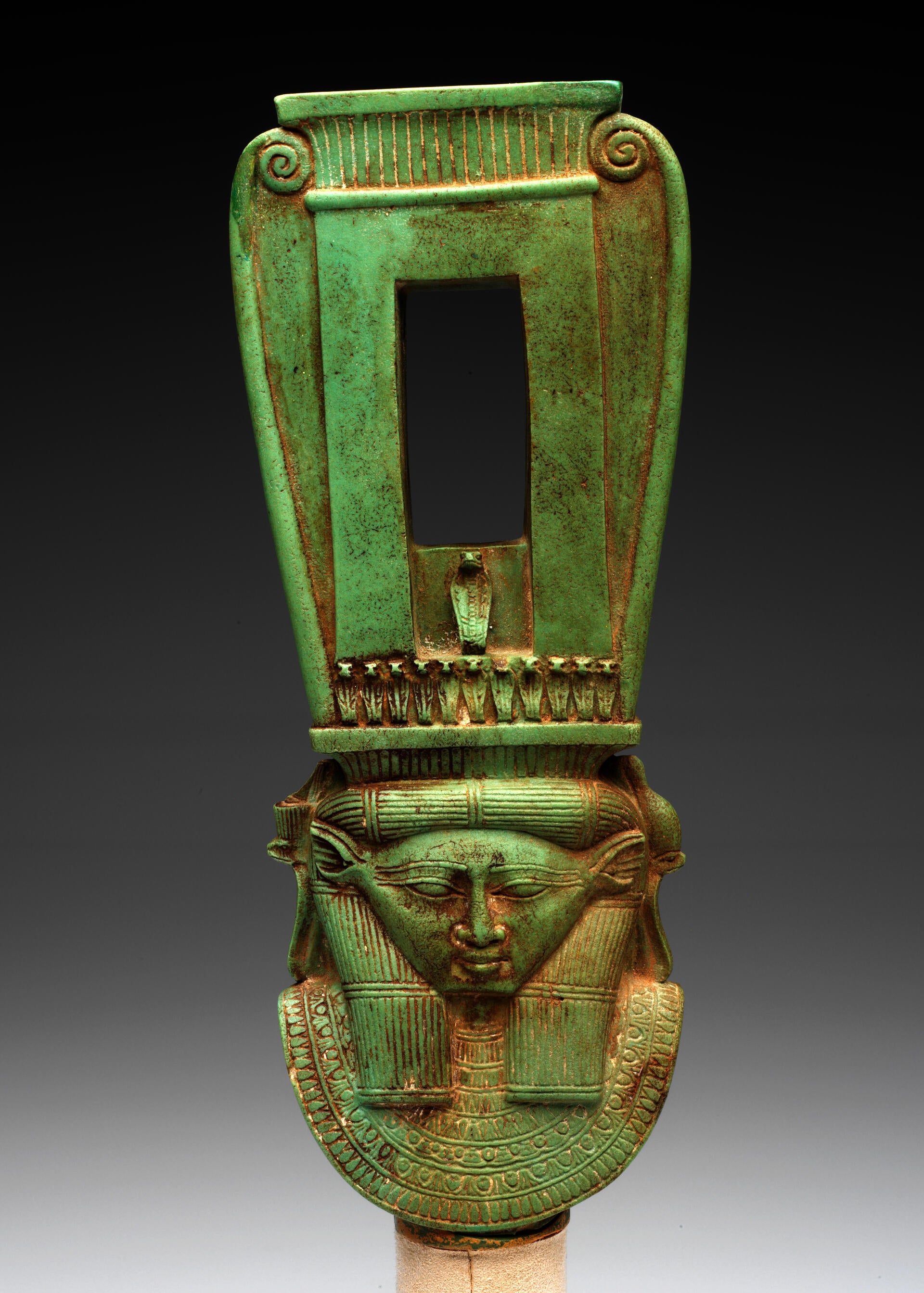 Front view of a long jade-green sculpture of a collared figure wearing a tall headdress, which has a rectangular opening and swirls on the outer edges forming a curved top.