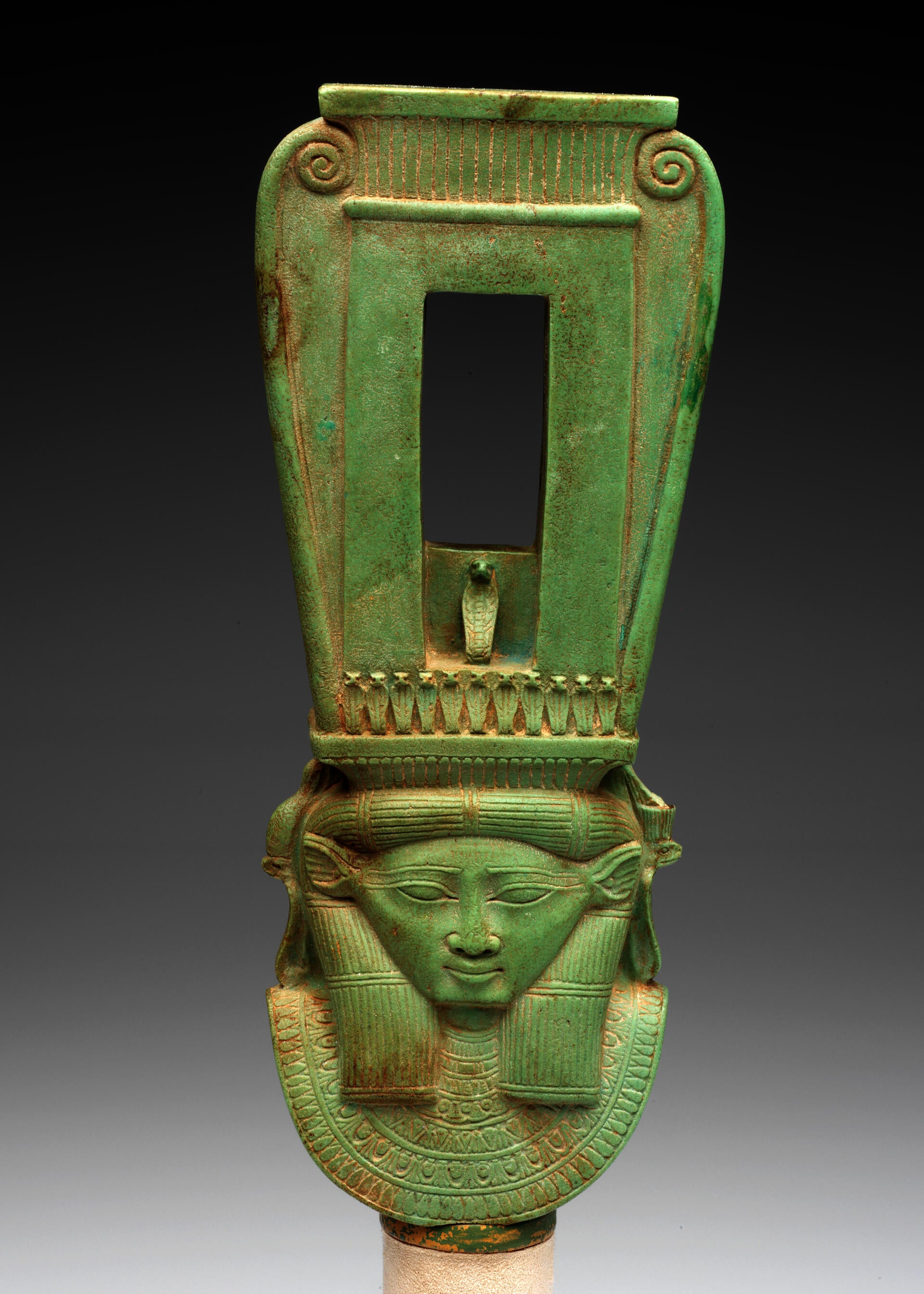 Front view of a long jade-green sculpture of a collared figure wearing a tall headdress, which has a rectangular opening and swirls on the outer edges forming a curved top.