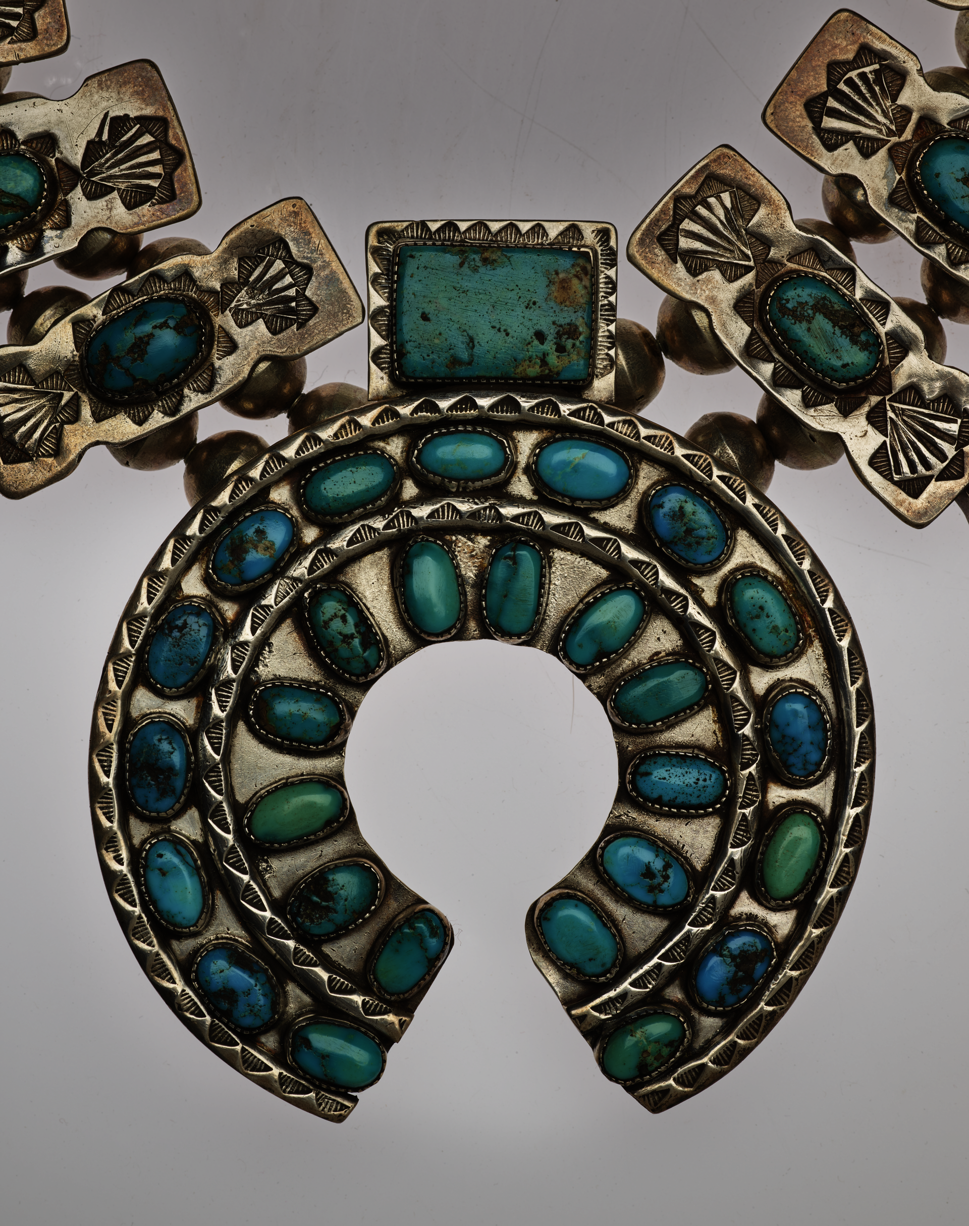 Detail-view of a horseshoe-shaped pendant of a turquoise and silver necklace. The turquoise stones are set in two concentric circles with decorative silver borders with a rectangular stone-setting atop it.