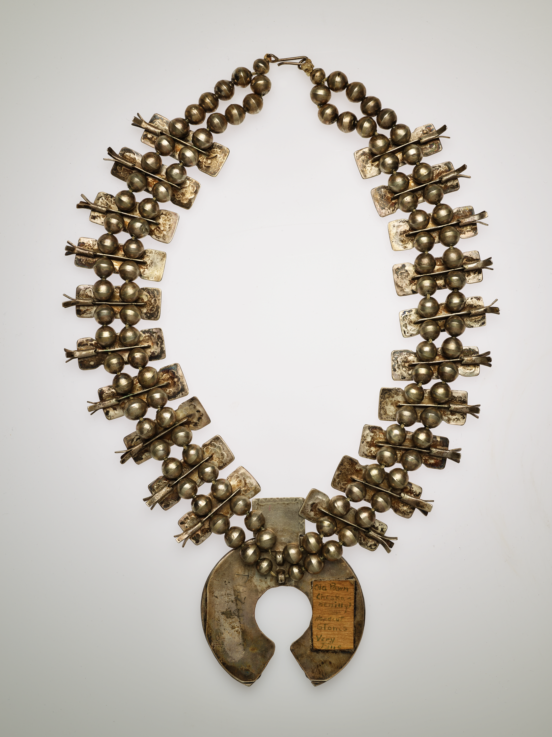 The back of a silver pendant necklace with two rows of silver beads throughout. On the central larger pendant there is a rectangular cream cloth on the right side of the semi circle pendant.
