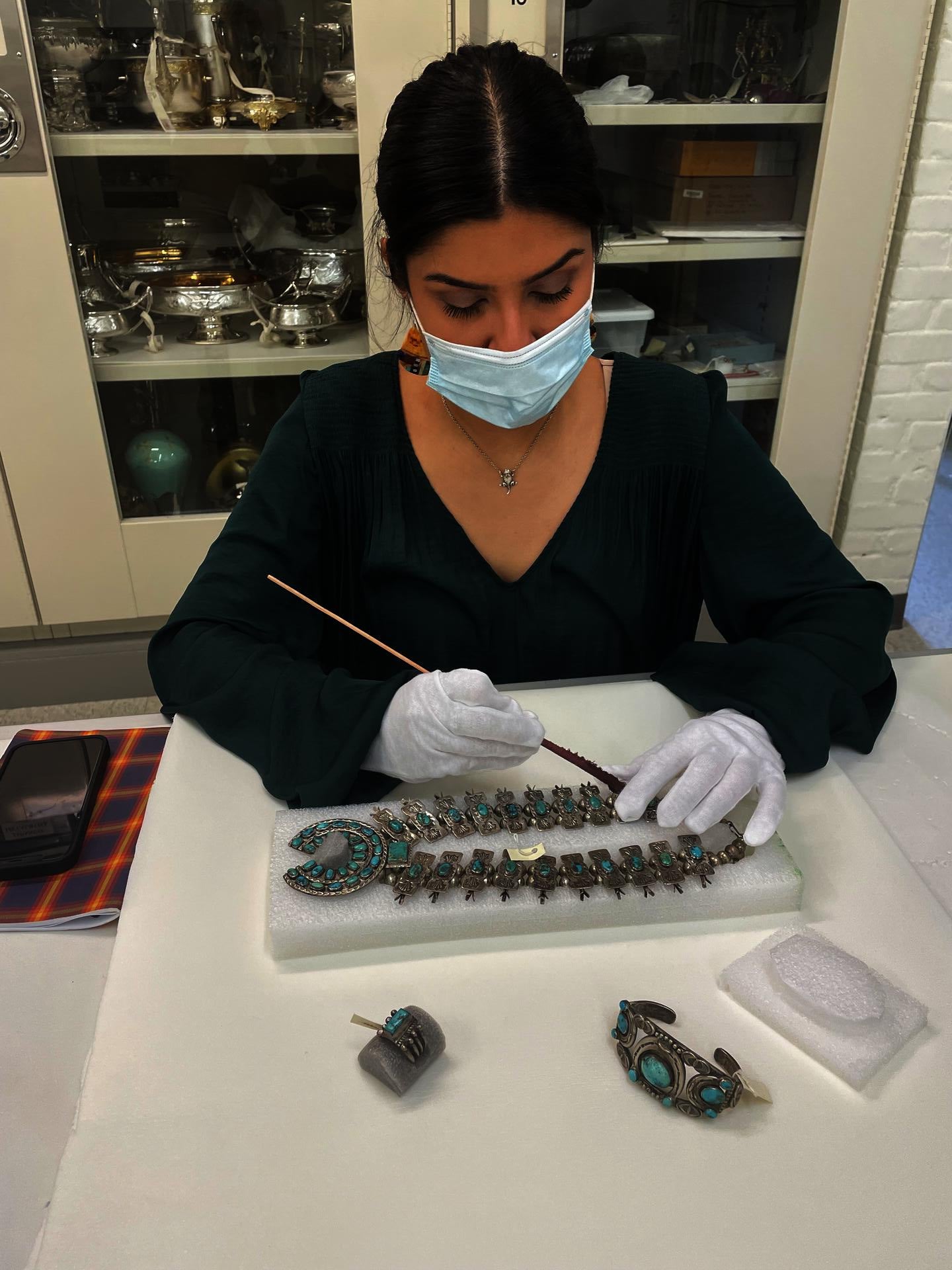 Picture of a person, wearing a mask and gloves, holding a long thin device to a silver and turquoise necklace laid in front of them on a foam rectangle.