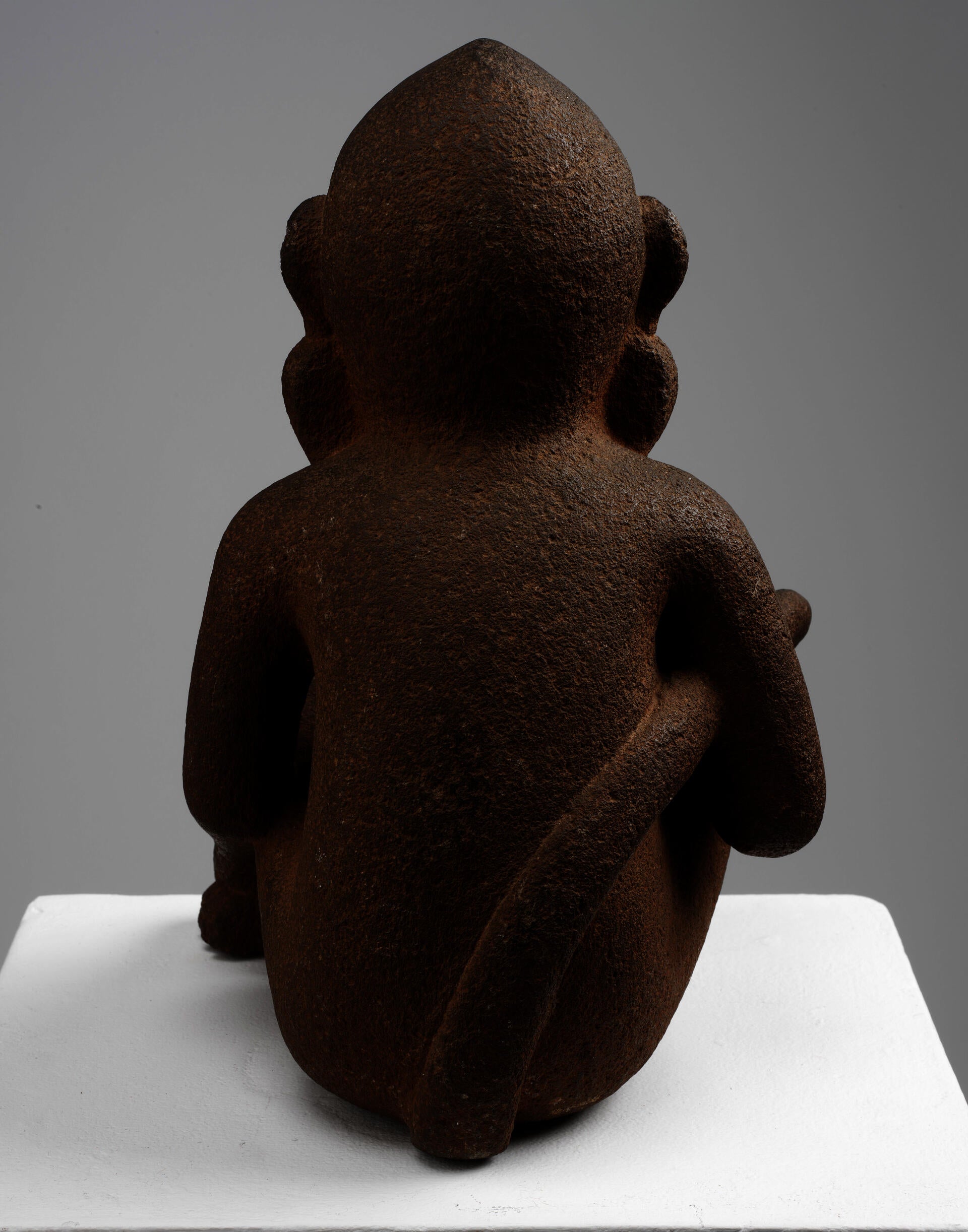 Back of a brown, porous-textured sculpture of a sitting monkey with a long tail held in its right arm. It is propped on a white rectangular platform.