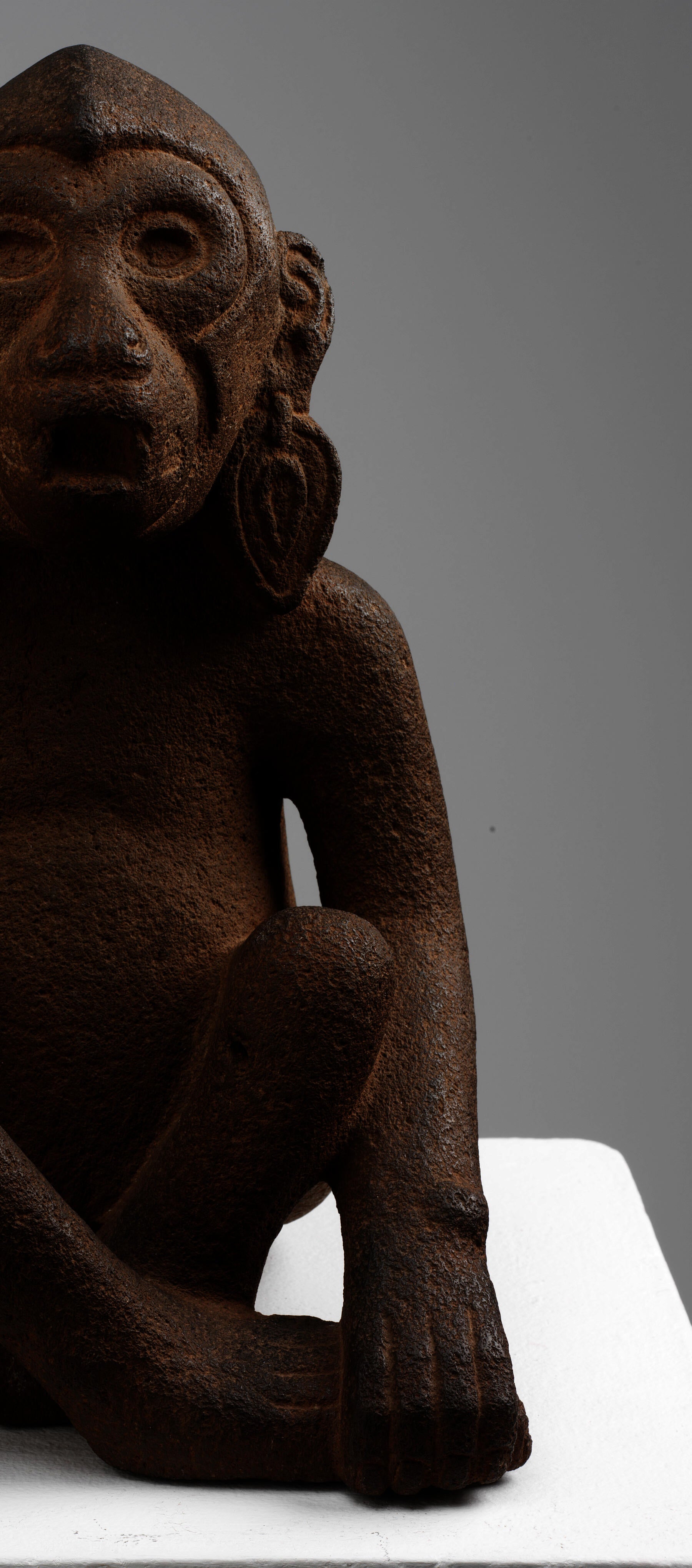 Cutoff view of the front right-side of a brown monkey sculpture sitting cross-legged with one hand gripping its foot. Its hands and toes are engraved to delineate toes and fingers.
