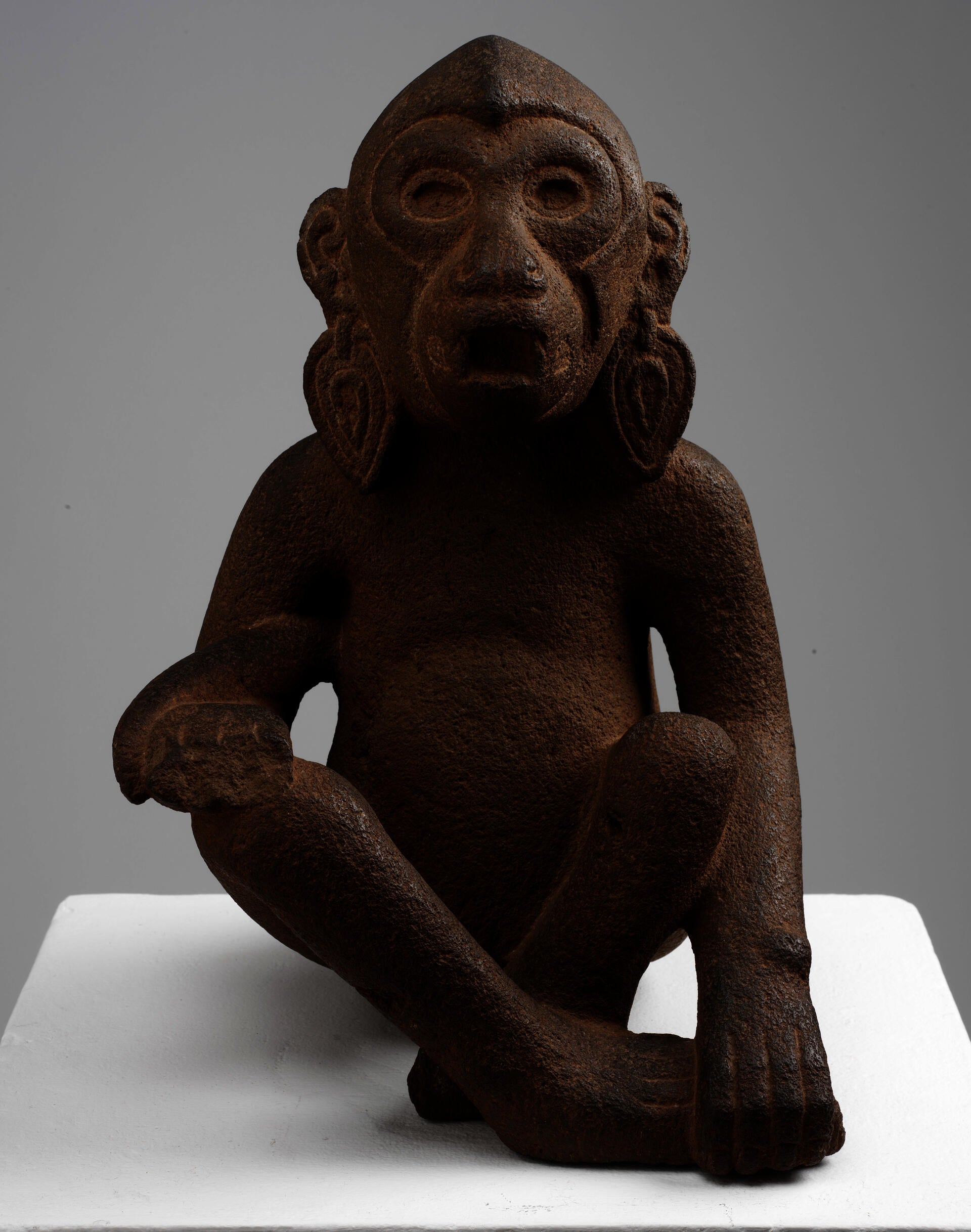 Front of a brown sculpture monkey wearing earrings. Its tail drapes over the left arm, and its right arm holds its left foot. Its eyes and mouth are open.