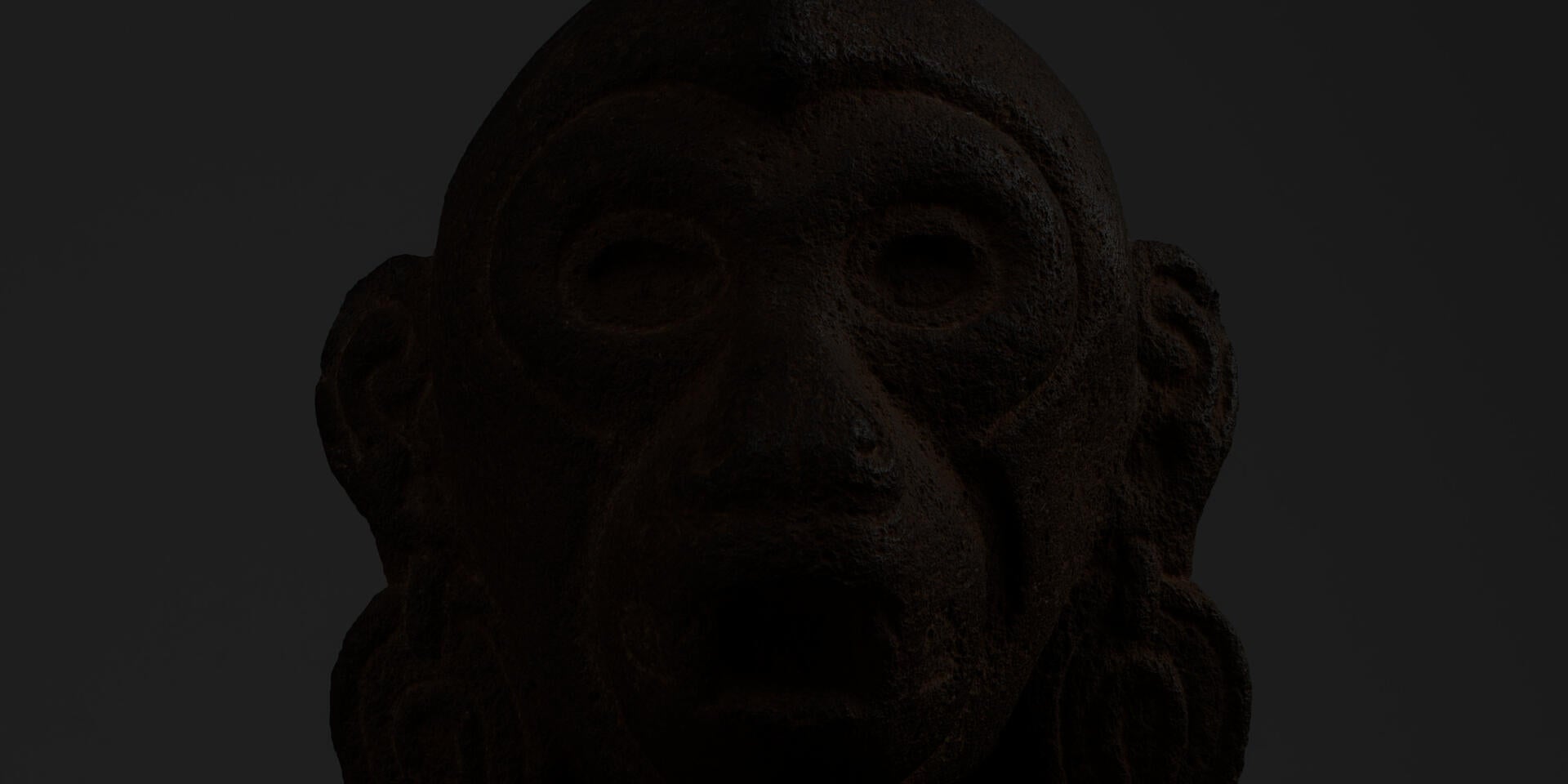 Detail of a dark brown, porous sculpture of a monkey head with a gray background. The monkey has wide open eyes, large earrings, and an open mouth.