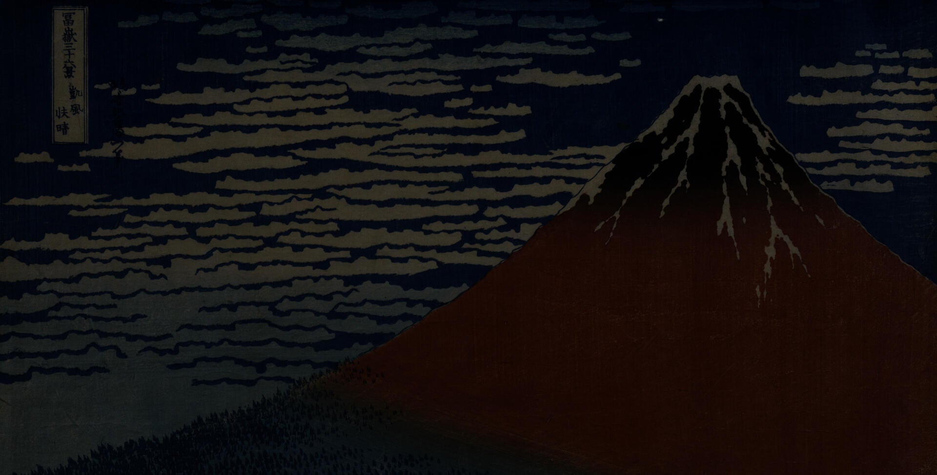 Woodblock print of a large mountain colored red and blue with white clouds and a blue sky behind it. Small trees sit at the base of the mountain. In the top left of the frame, Japanese text is in a white rectangle.