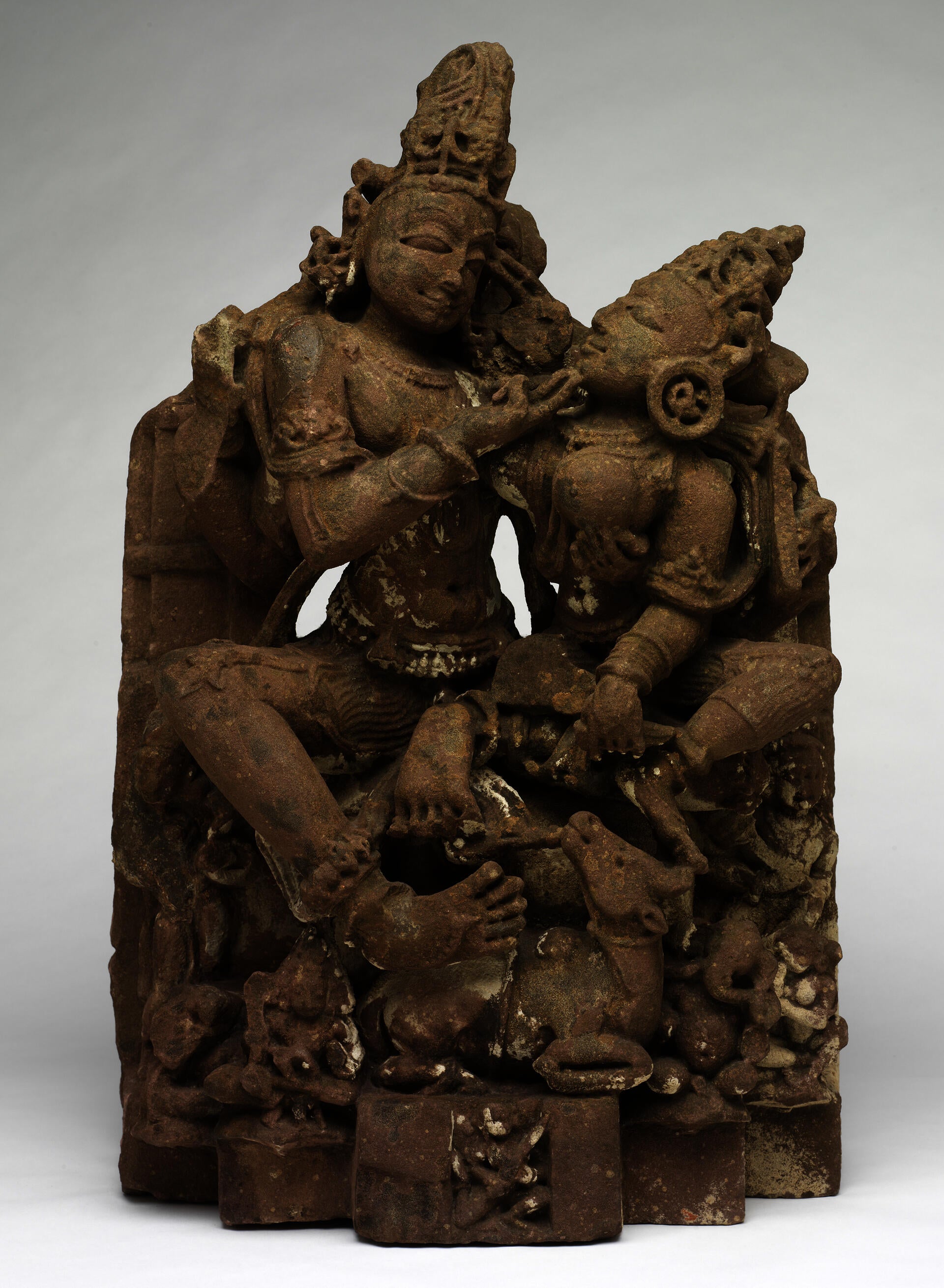 Brown sculpture of two figures caressing each other. Both figures wear headdresses and minimal clothing. Various animals watch from below, and they are all situated on a platform.