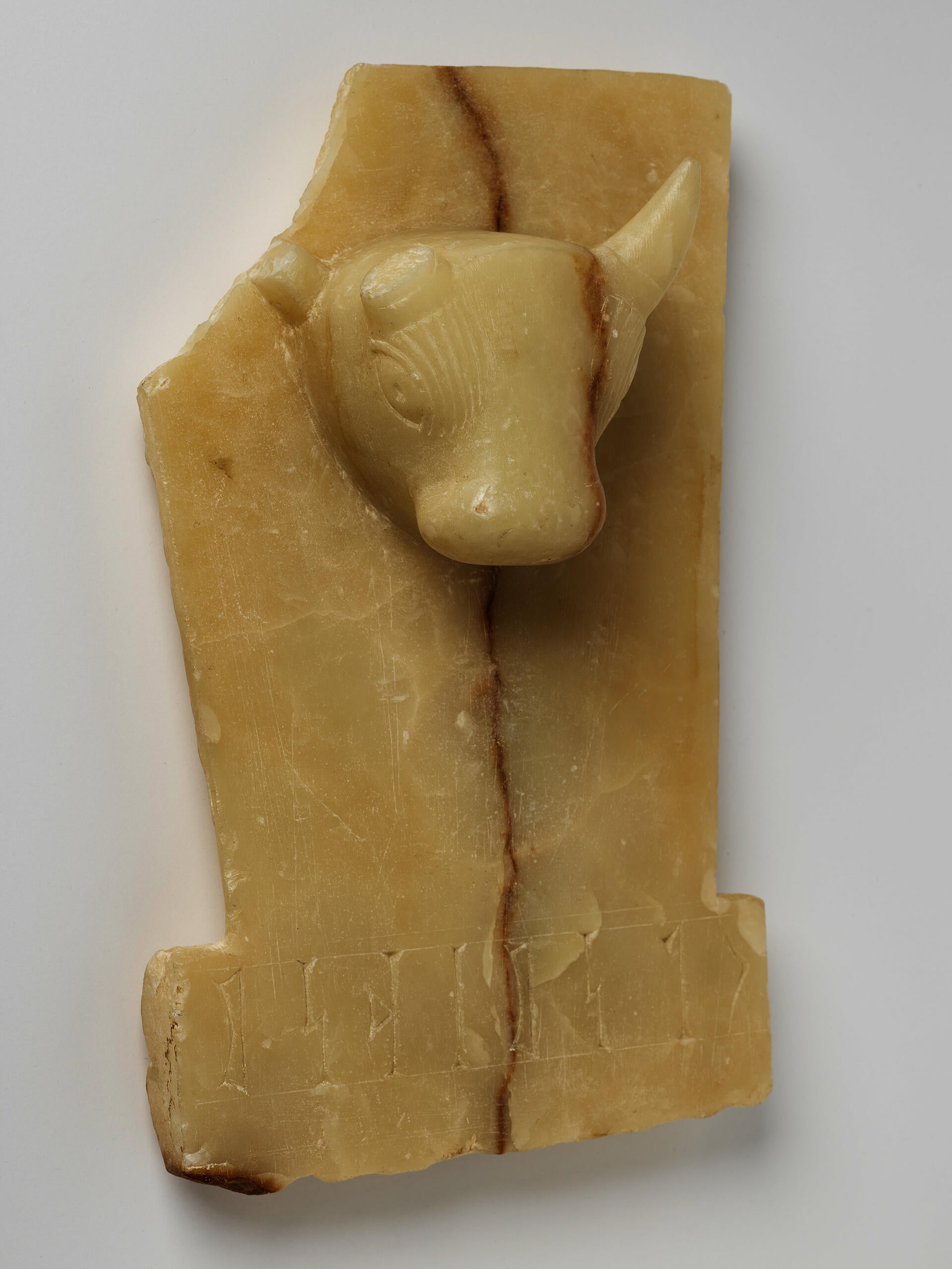 Cream rectangular stone with a sculpted bull’s head emerging from the upper center. Its upper left is broken off. A brown crack runs vertically down the center.