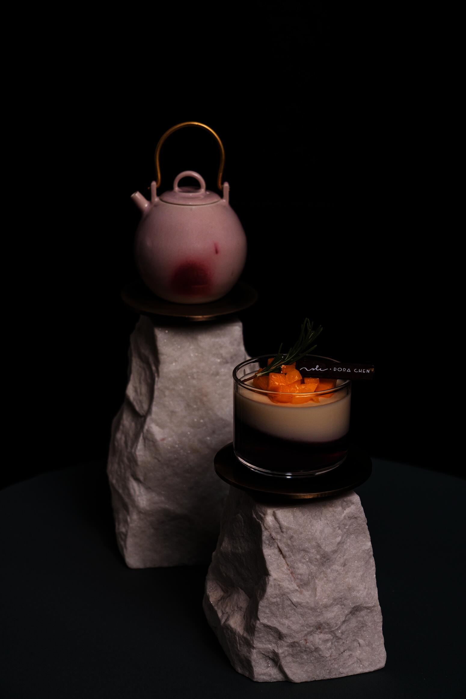 A pink glazed ceramic teapot with a brass handle and a panna cotta dessert in a small glass cup is displayed on two stone stands.