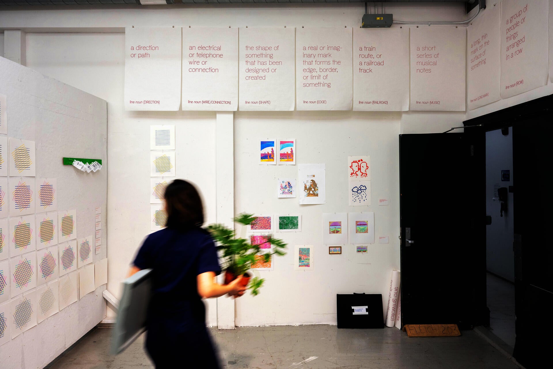 A room with a variety of small-scale works pinned to white walls. A person in motion blur walks in front of it, carrying a plant.