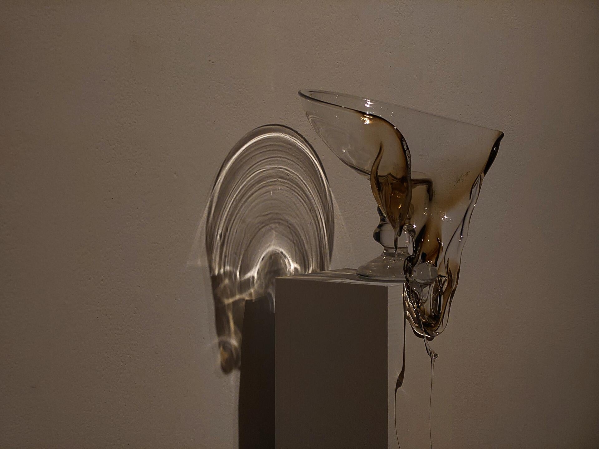 A slumped hand-blown goblet receiving a spotlight, casting a haloed shadow on the wall.