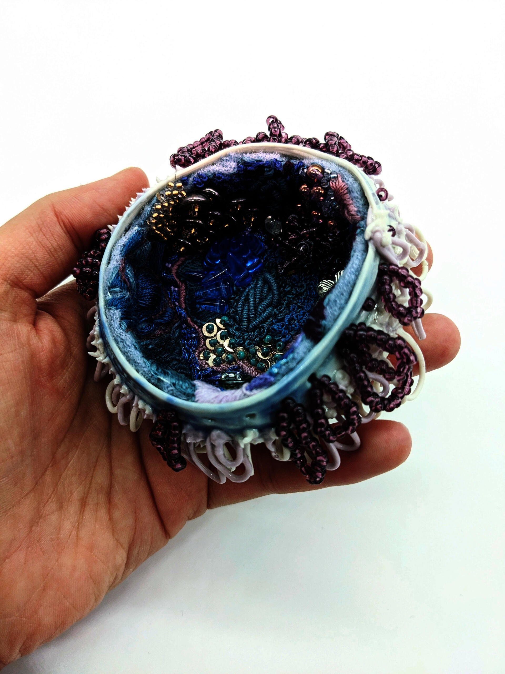 hand holding small porcelain vessel lined with beads and embroidery