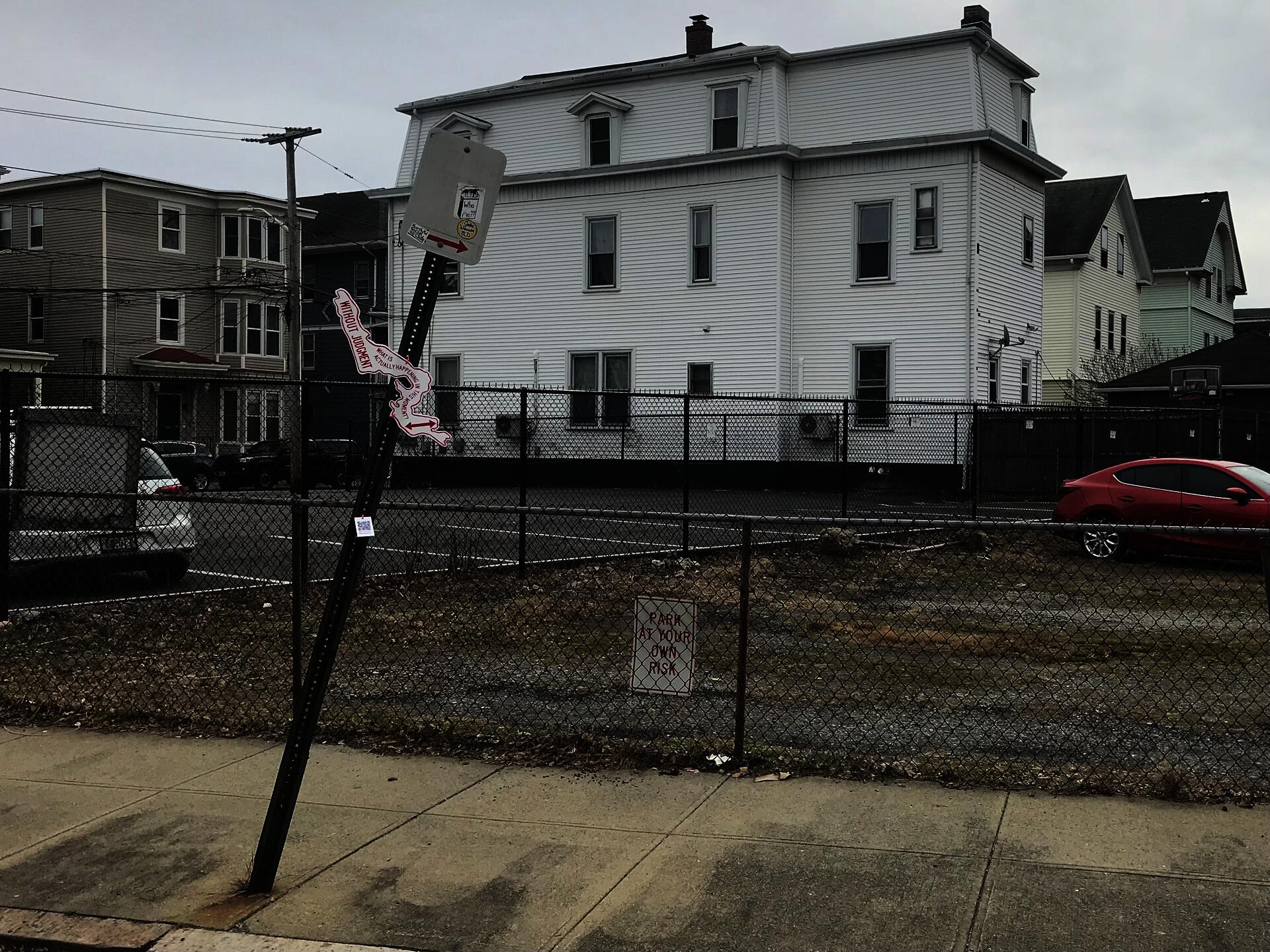 An organically-shaped red and white sign posted on a “No Parking” sign post, which reads, “Without judgment, what is actually happening in this moment?”