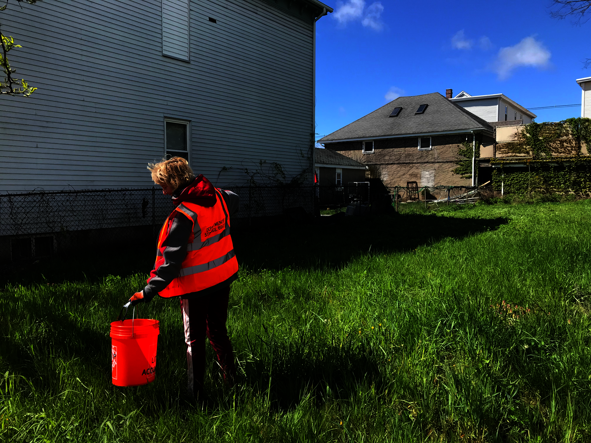 A woman in an orange vest with “Department of Strange Beauty” on the back uses a trash grabber to drop litter into an orange bucket.