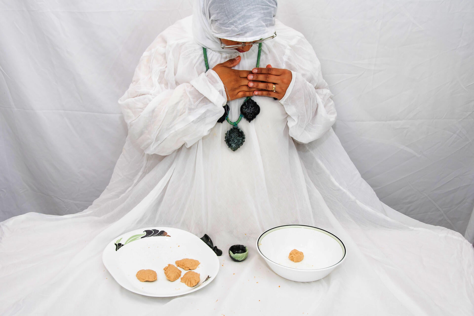 Figure with white garment draped over a table wearing a necklace with soapstone molds pressing mishti against her chest .