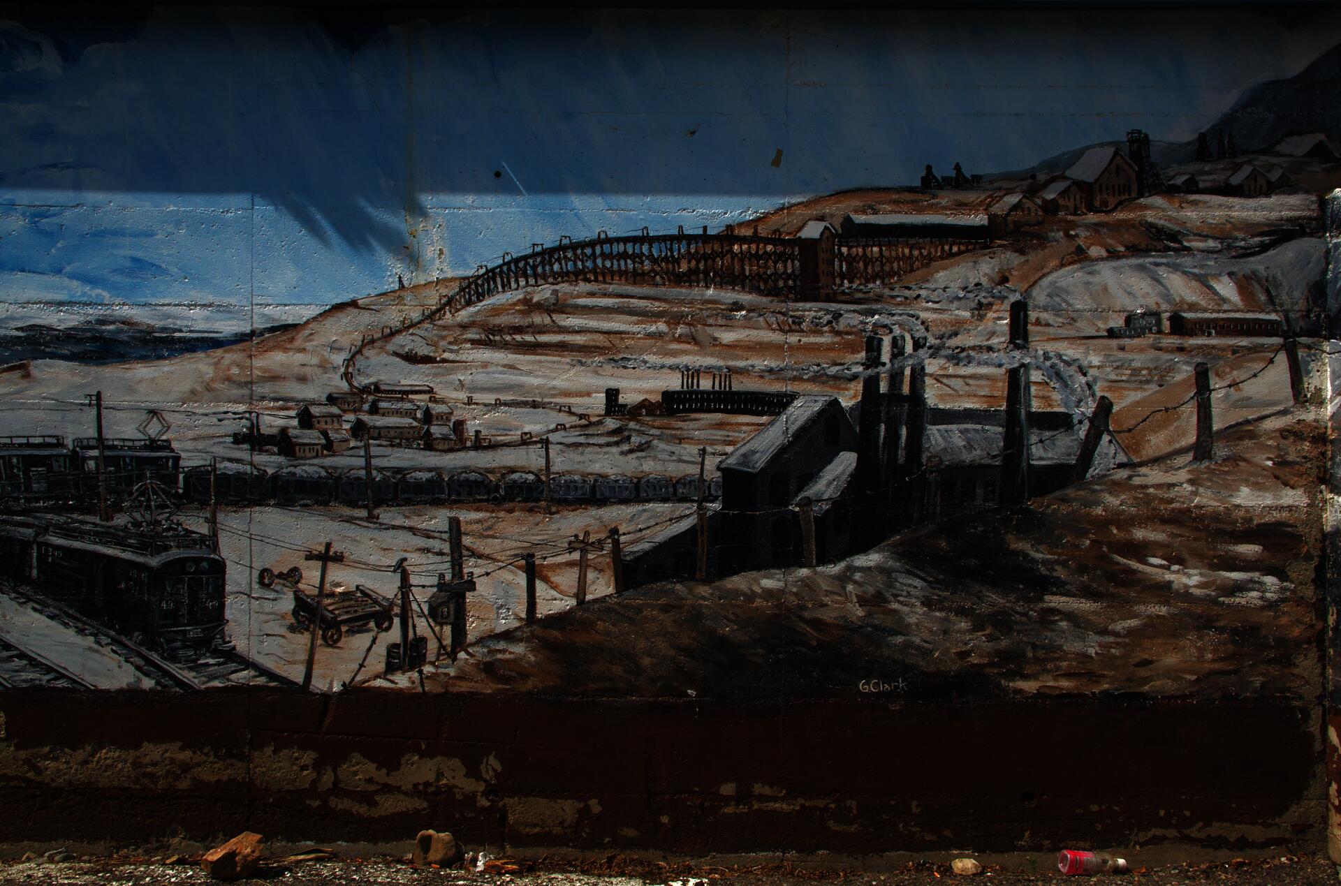 Photograph of landscape mural depicting the settler copper industry in Butte, Montana in the early twentieth century. A shadow of a plant is visible against the mural’s blue-painted sky. In front of the mural, pieces of trash lie strewn about.