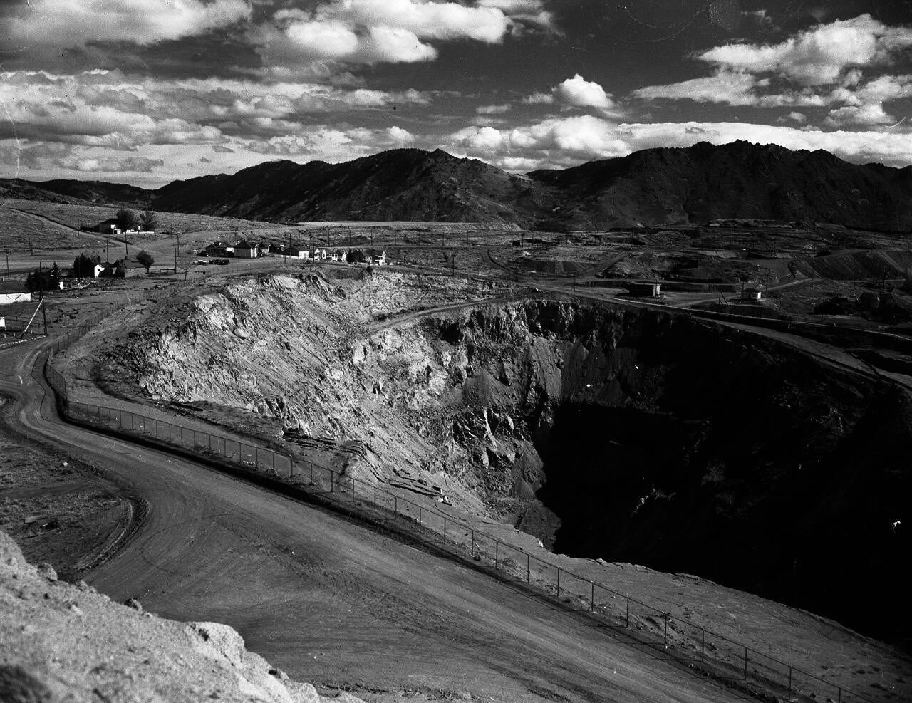 An open-pit copper mine with a caved-in wall gapes open in the foreground. A shadow blankets the southern rim of the pit. The heavily mined ground beyond the pit slopes up to meet the Continental Divide, from which water flows in two directions. Here is the source of Silver Bow Creek, draining into the Pacific Ocean.