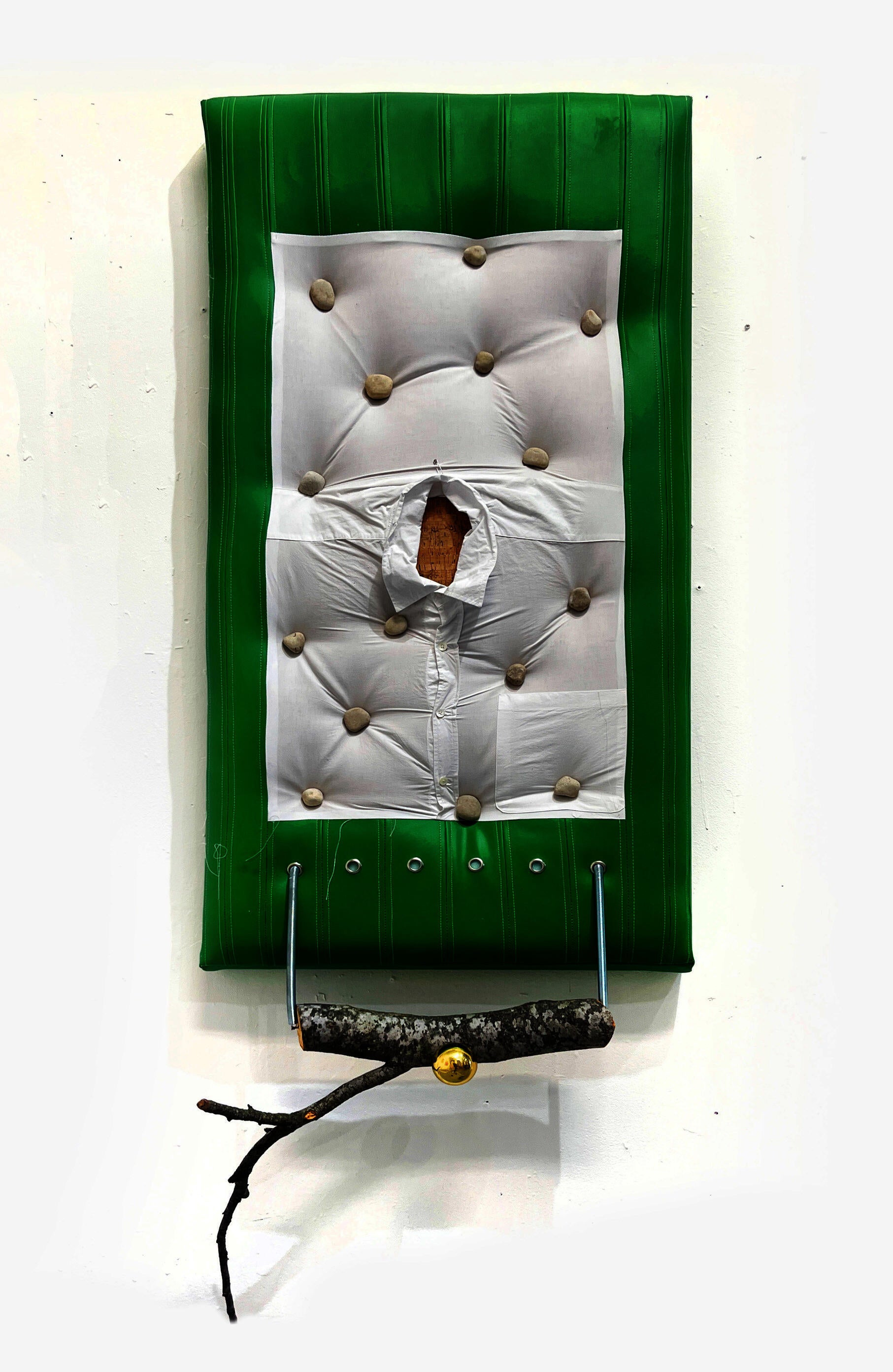 a painting, upholstered in green and adorned with a button down shirt and smooth stones holds a severed tree-limb from a pair of springs