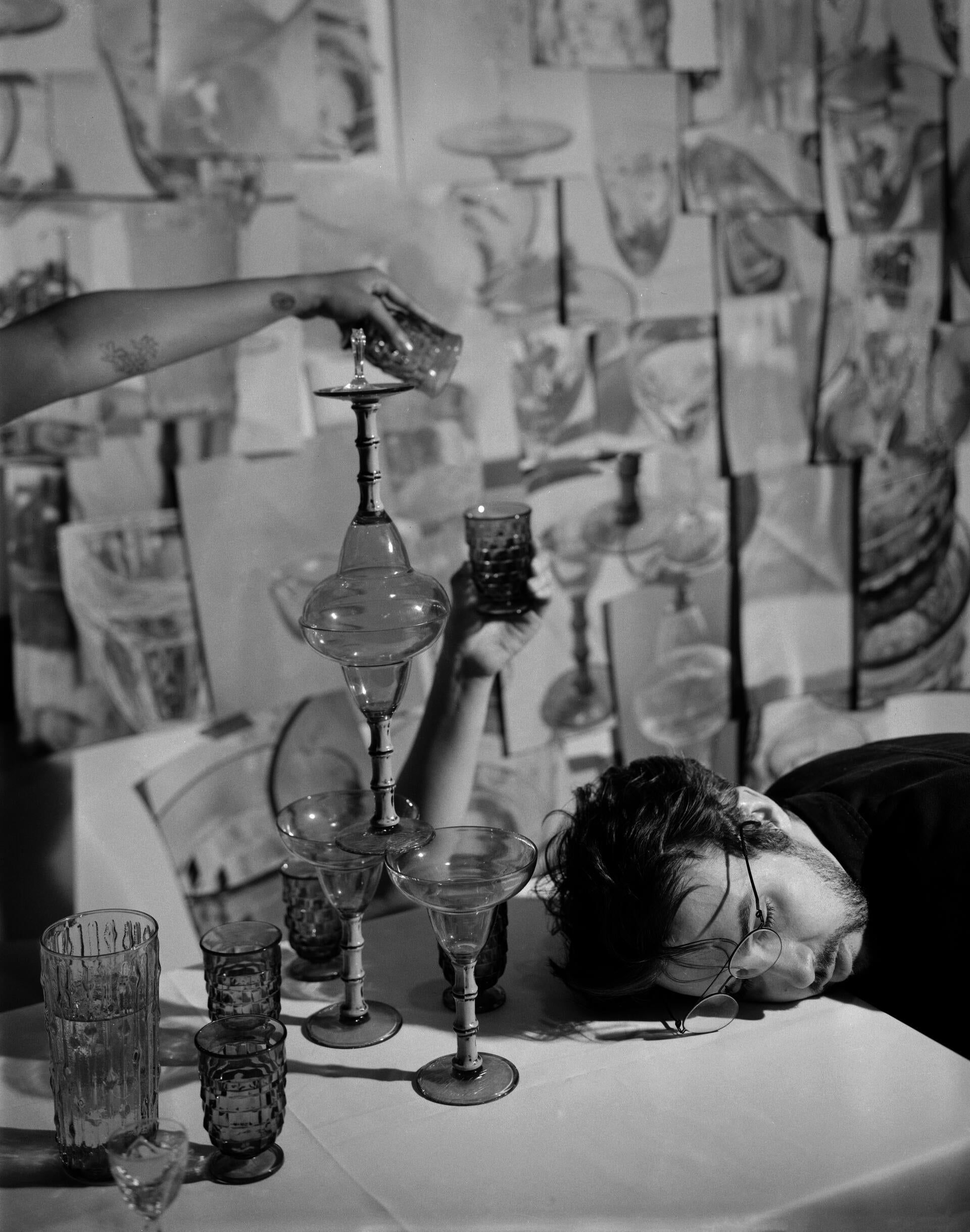 A man is lying on the table full of glasses, while two hands of others are holding glasses up in the air.