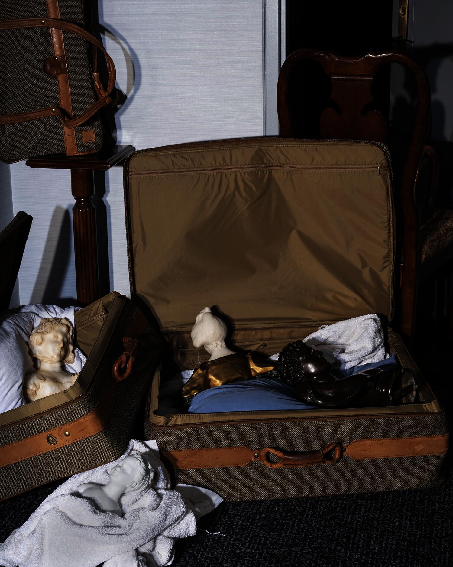 Color pigment print of busts in and around open suitcases.
