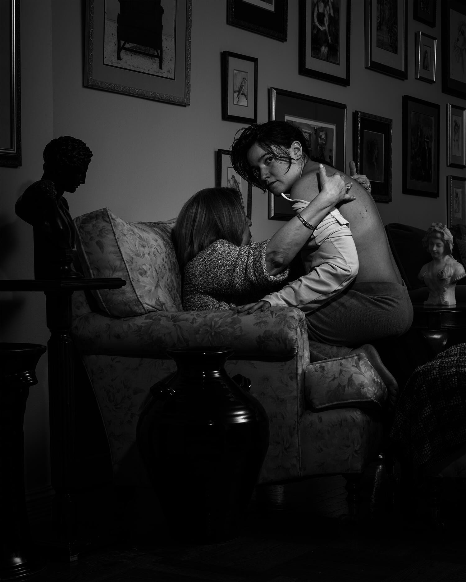 Black and white pigment print of young adult sitting on older adult's lap surrounded by framed images, vases and busts.