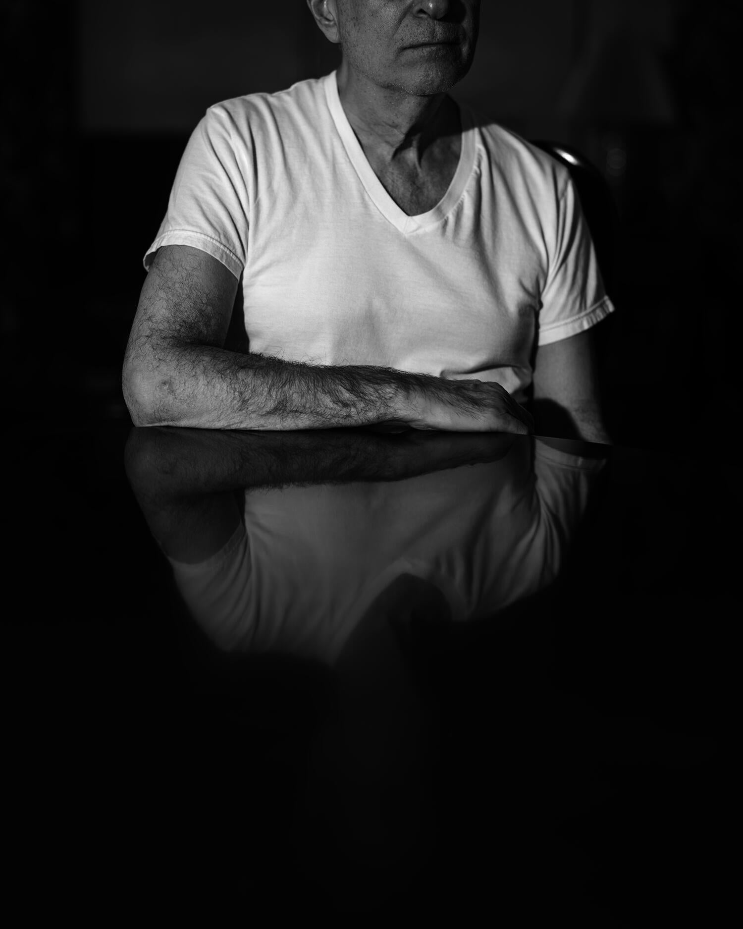 Black and white pigment print of man sitting with his obscured image reflected in dining room table.
