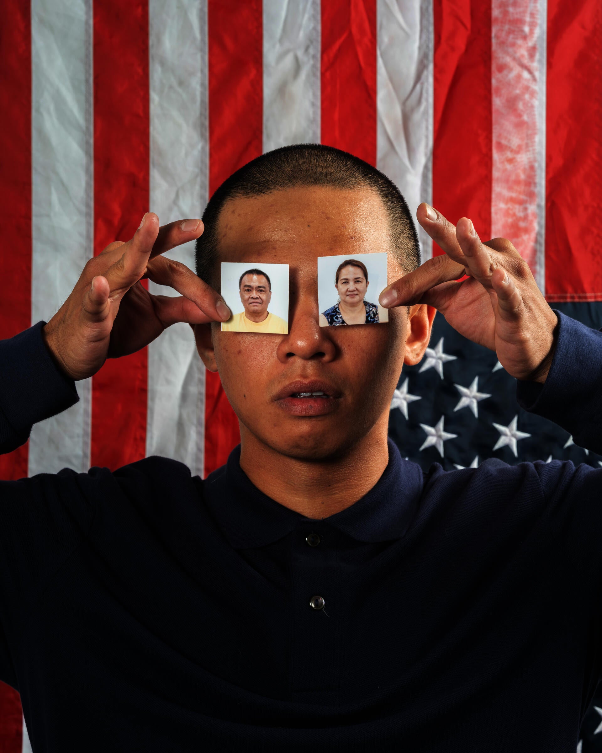A person holds up two passport photos, one in each hand, covering their eyes with it against an American Flag