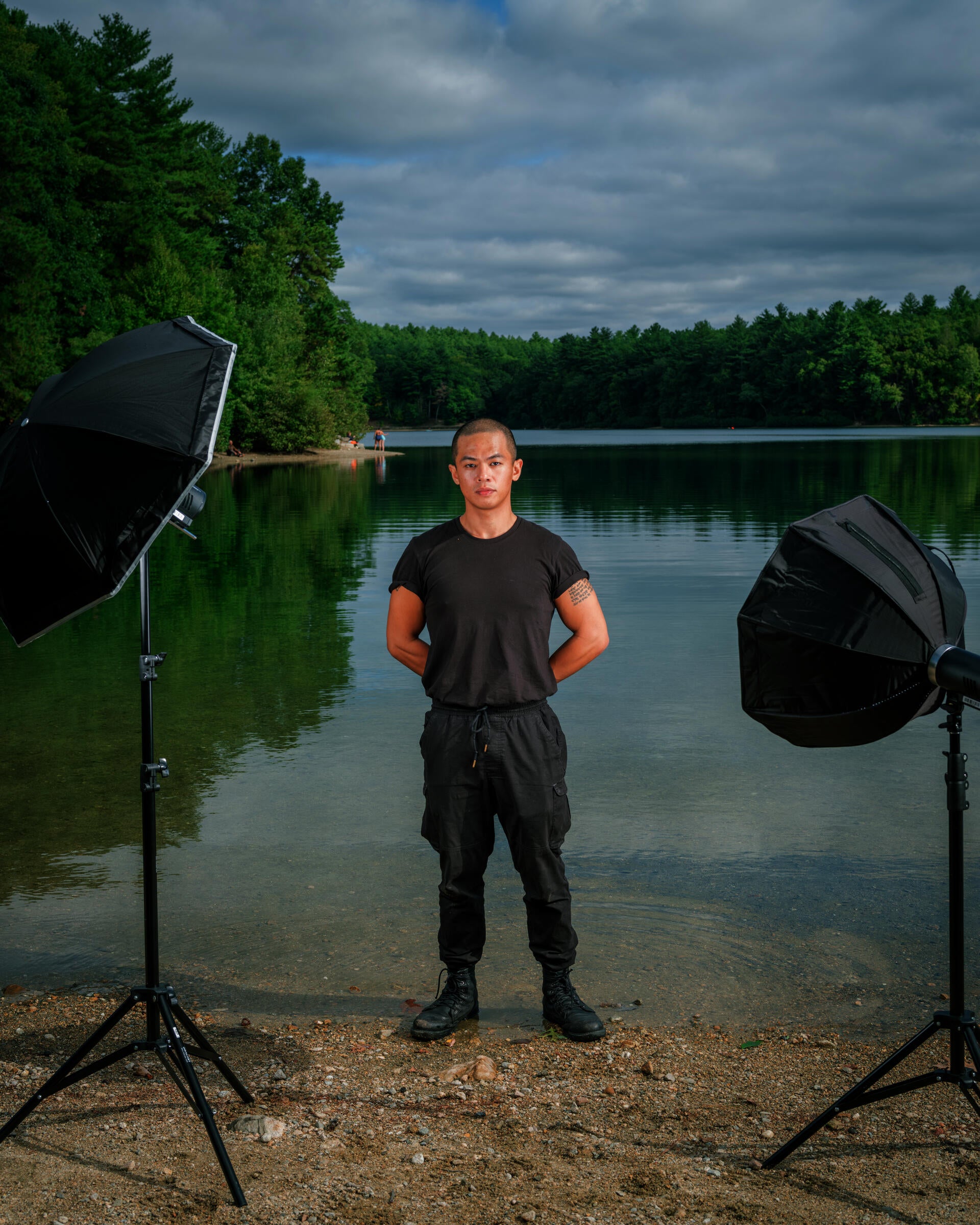 A man wearing all black standings shoulder-width apart at the shore of lake. Two flash strobes frame him.