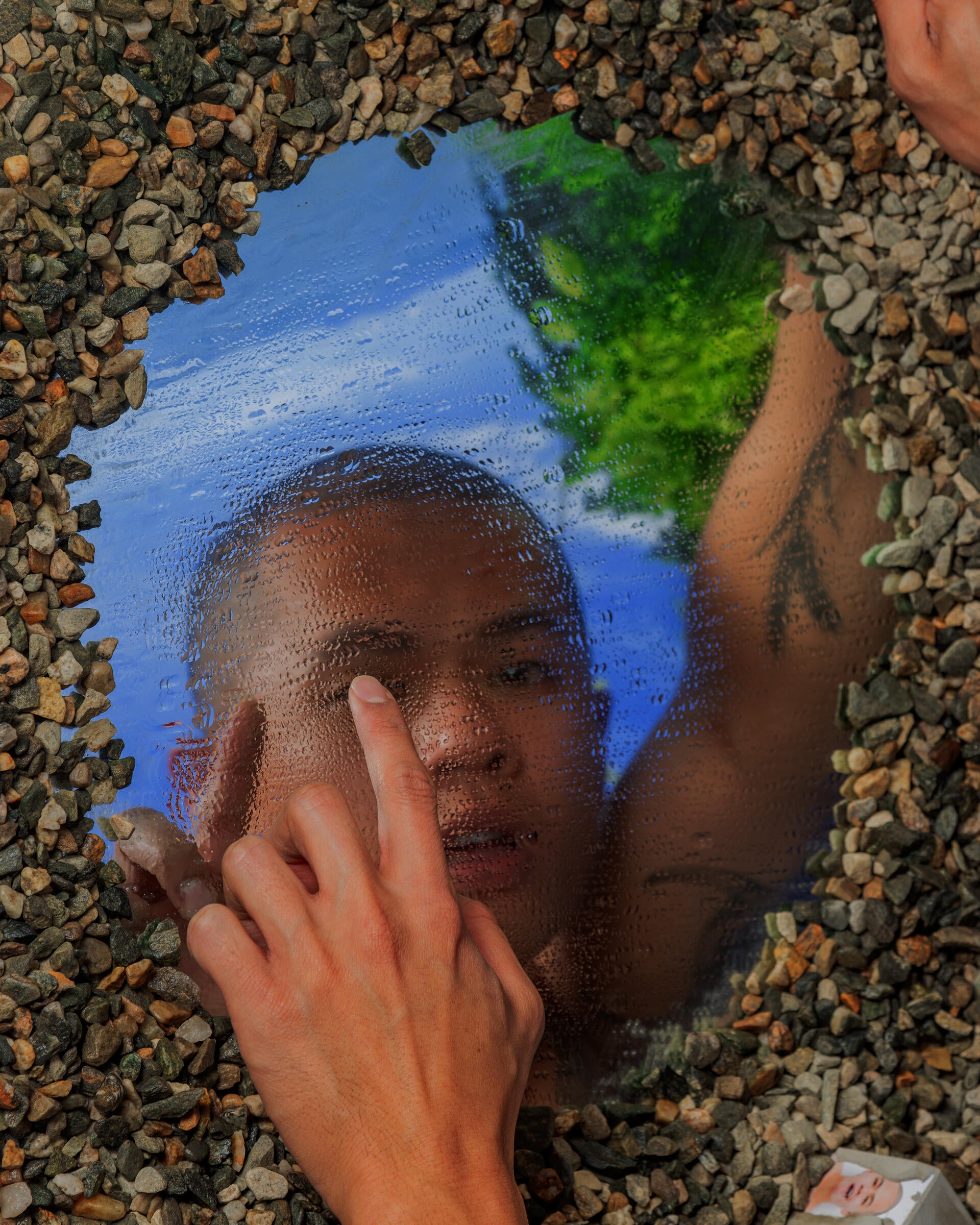 A man is touching a circular pool showing his reflection, the pool is surrounded by gravel.
